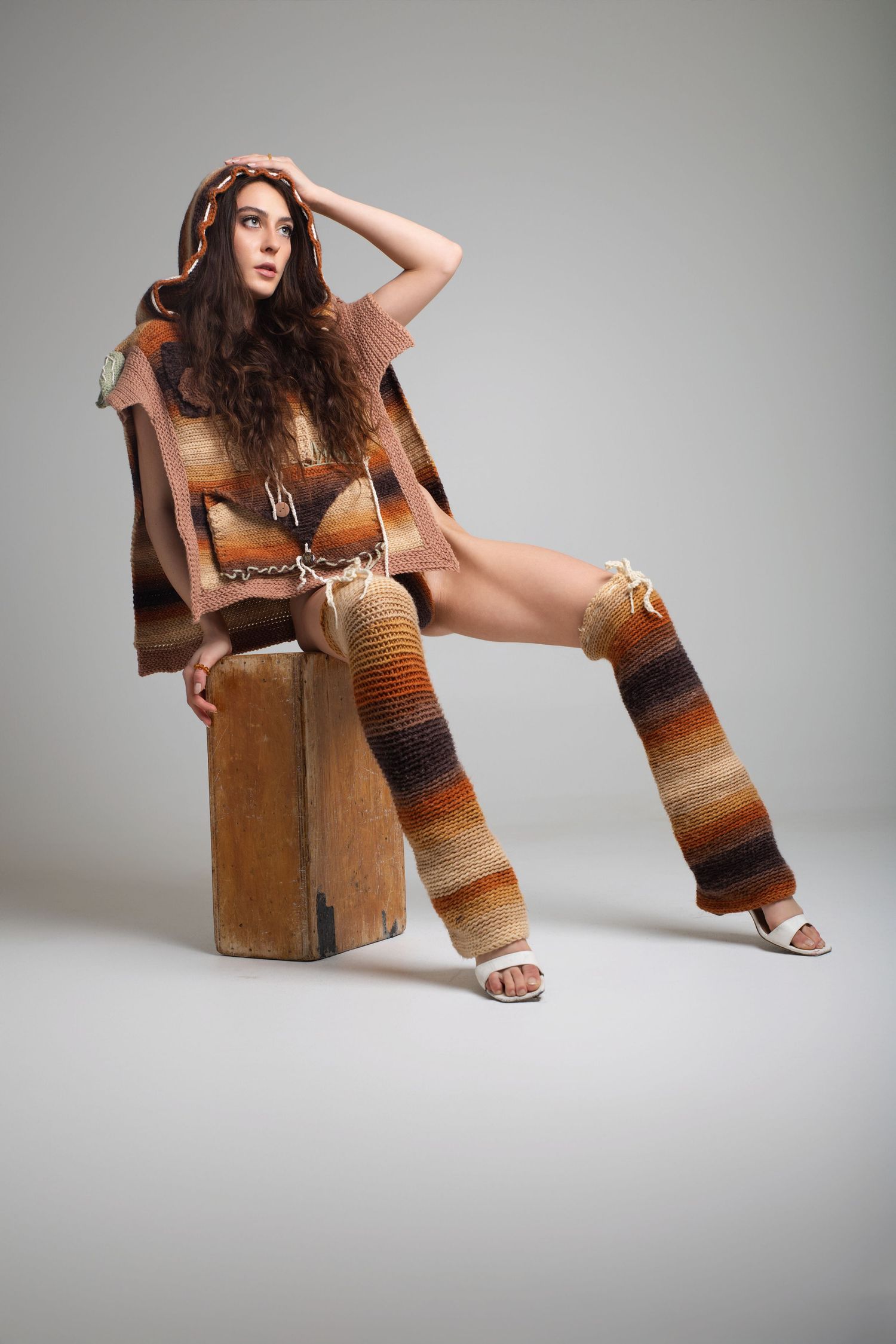 Brunette woman seated on wooden block wearing a brown striped hooded knit outfit with leg warmers.
