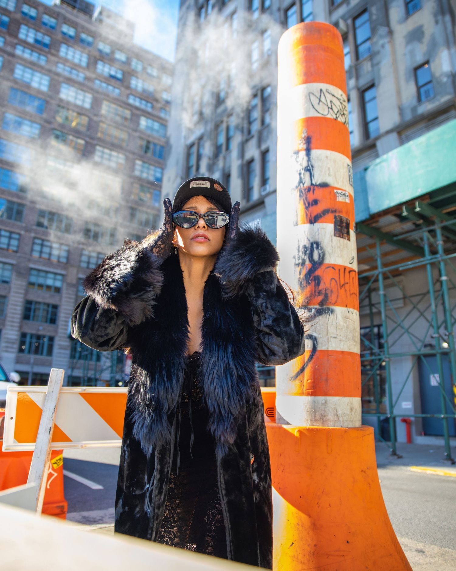 Woman in black fur coat and sunglasses posing by orange construction barrier.