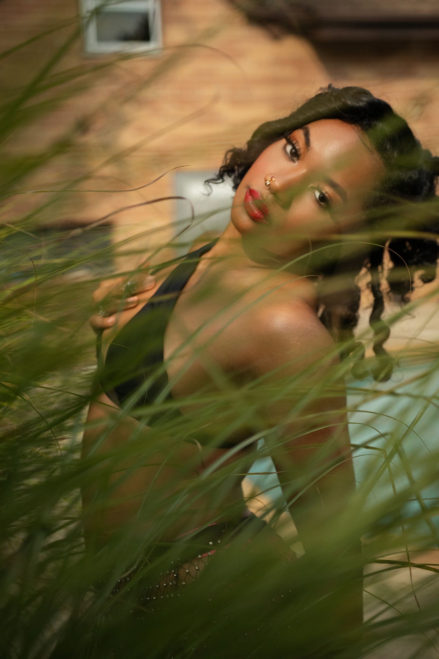 Woman with red lips peering through tall green grass in warm outdoor setting.