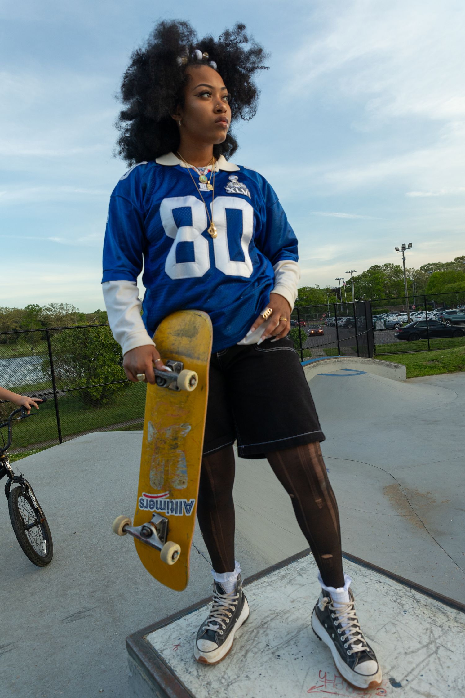 Young Black woman holding a yellow skateboard at a skate park, wearing a blue number 80 football jersey and black shorts.