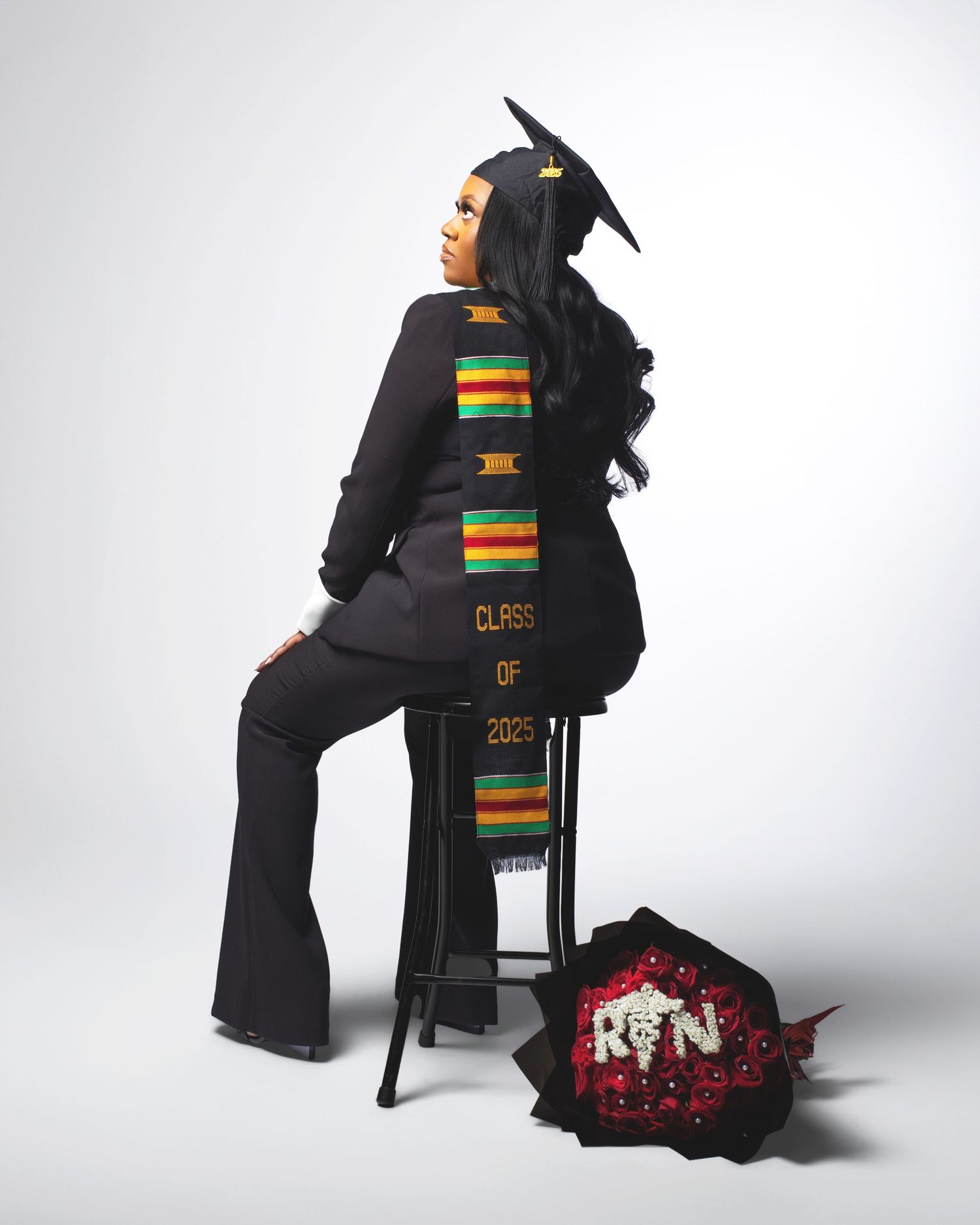 Graduate seated on stool wearing Class of 2025 RN kente stole and cap with floral bouquet.