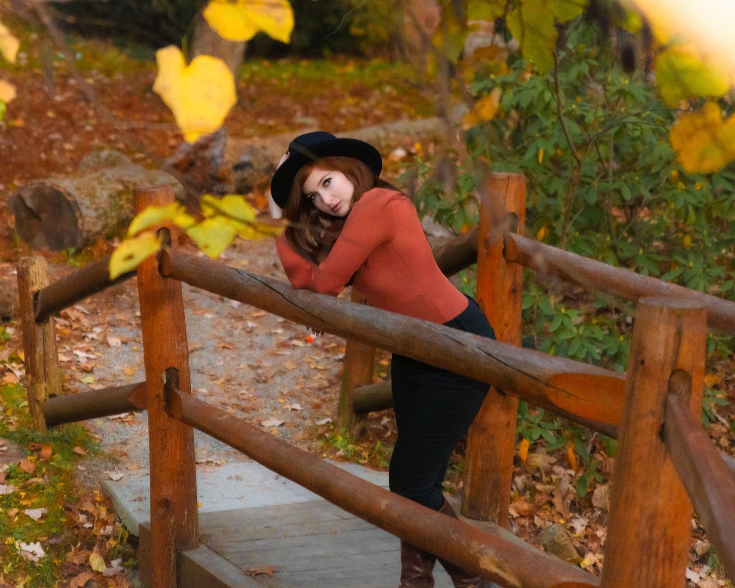 Young woman in orange top and black hat leans on rustic wooden bridge in fall park.