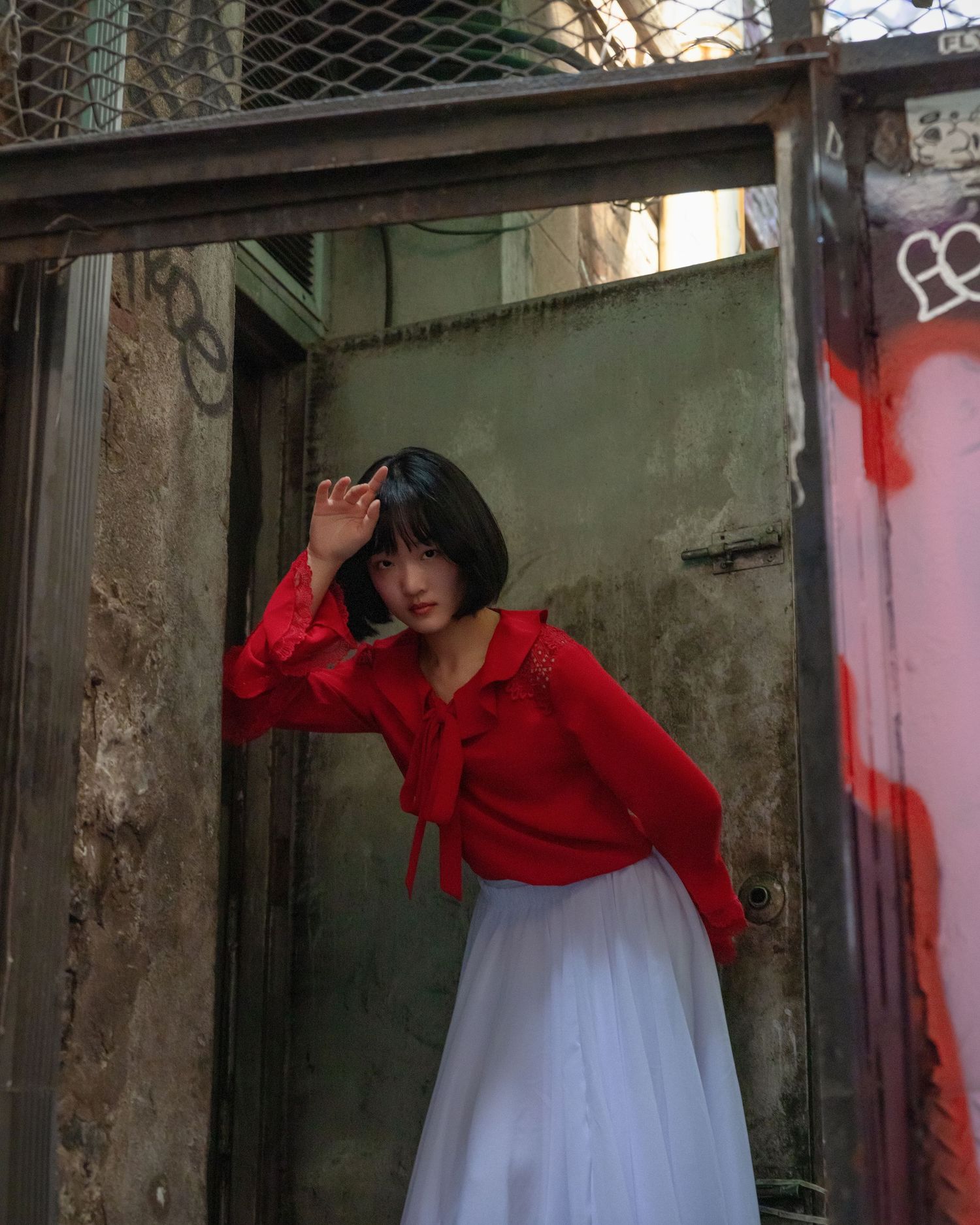 Young woman in red blouse and white skirt posing in urban alleyway with graffiti walls.