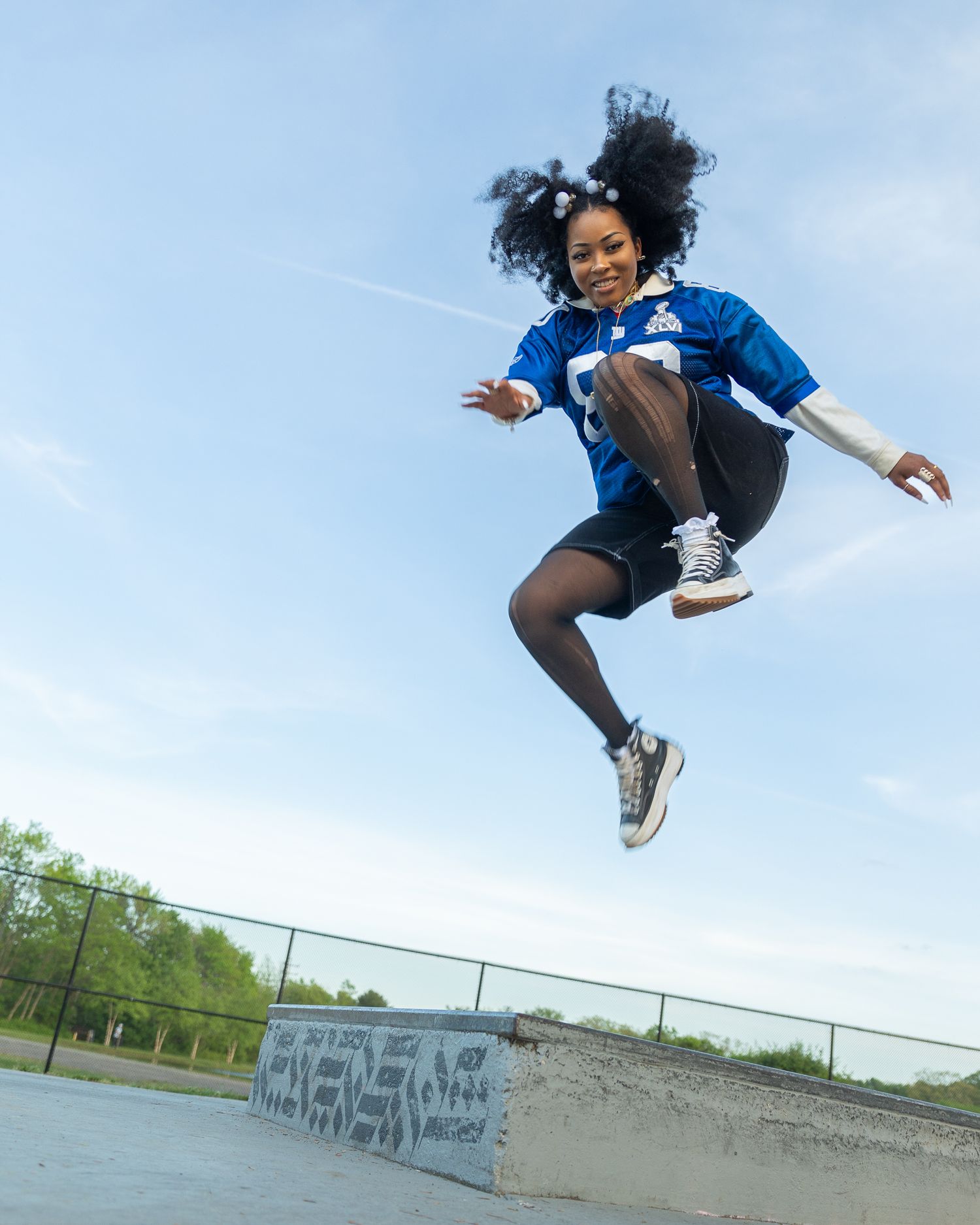 Energetic young woman jumping high at outdoor skate park wearing blue jersey and sneakers.