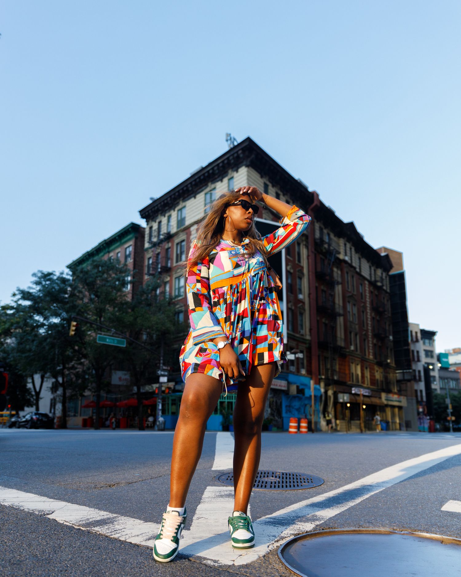 Fashion model in vibrant multicolor dress poses at NYC intersection under clear blue sky.