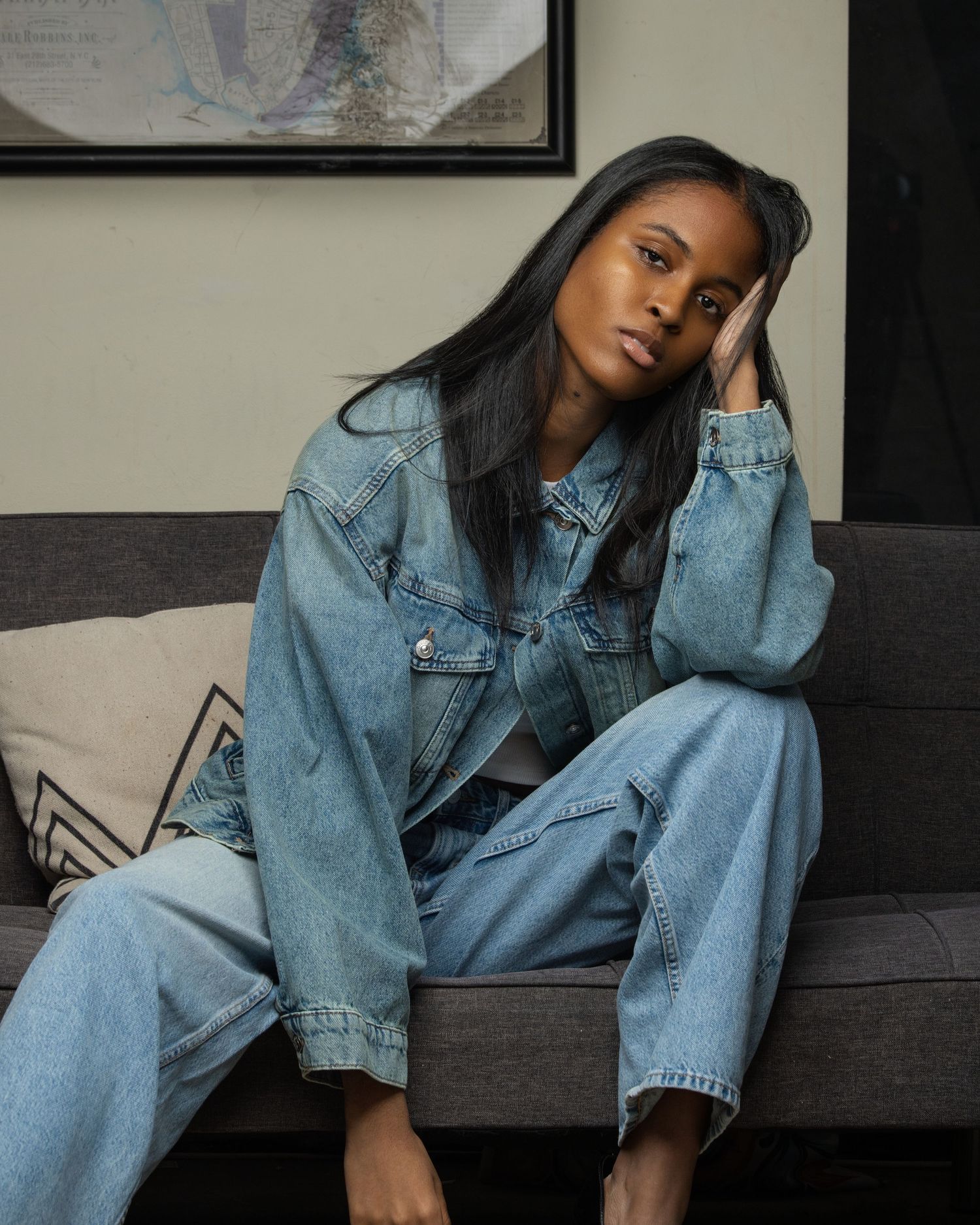 Young Black woman in denim jacket and wide-leg jeans sitting casually on gray sofa in relaxed pose.
