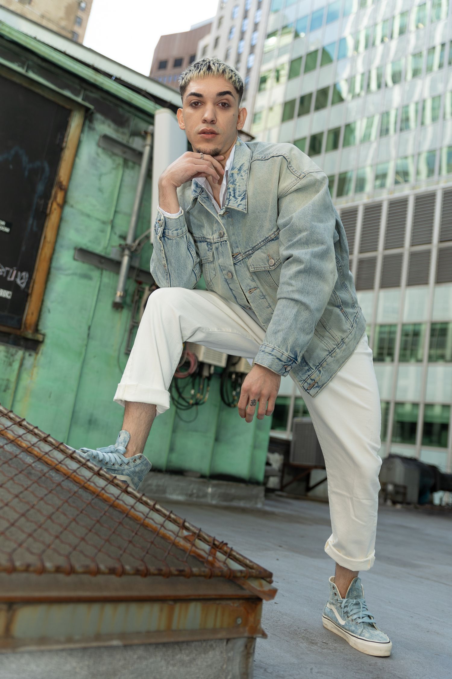 Young man in denim jacket and white pants posing on urban rooftop with city buildings behind.