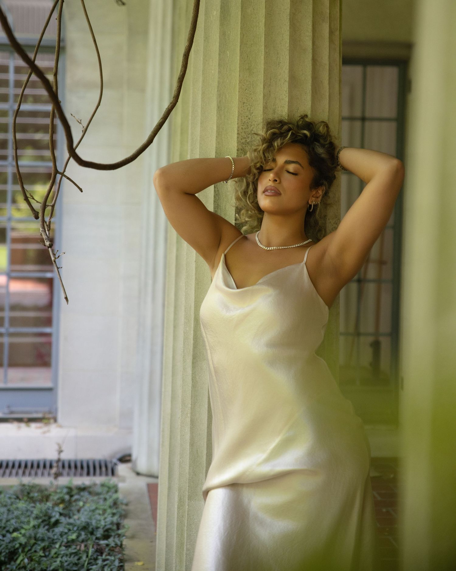 Woman in ivory satin slip dress with pearl necklace posing with hands raised near draped curtains and vines.