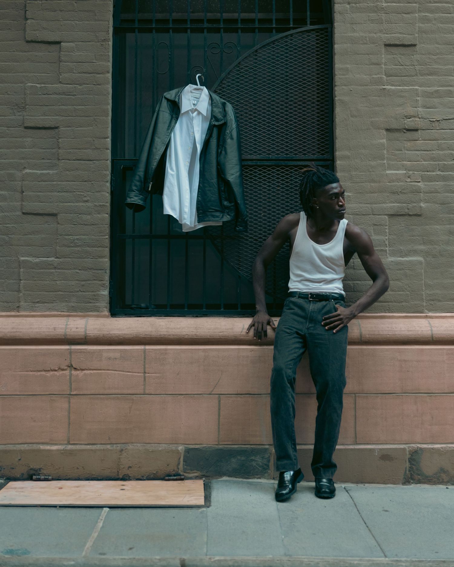 Young Black man in white tank top and dark jeans leaning against brick wall with jacket on hanger.