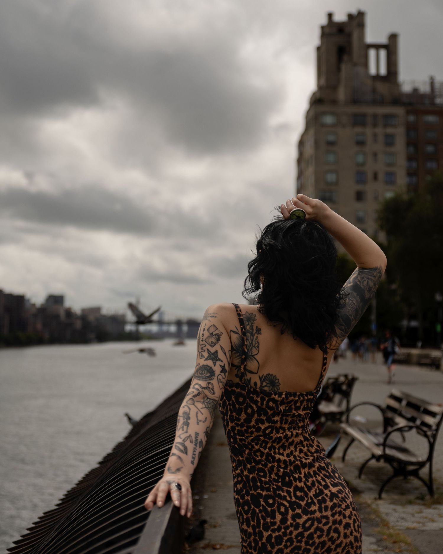 Tattooed woman in leopard print dress overlooking a river with a cloudy urban skyline backdrop.