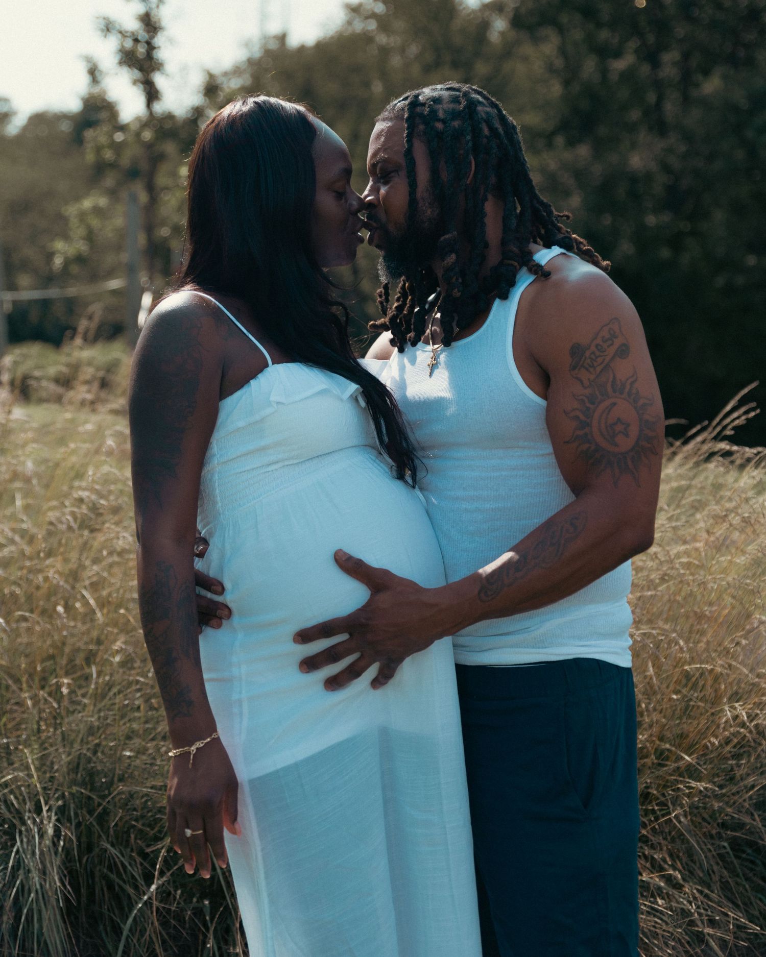 Expecting Black couple in white outfits sharing tender moment in golden field during maternity shoot.