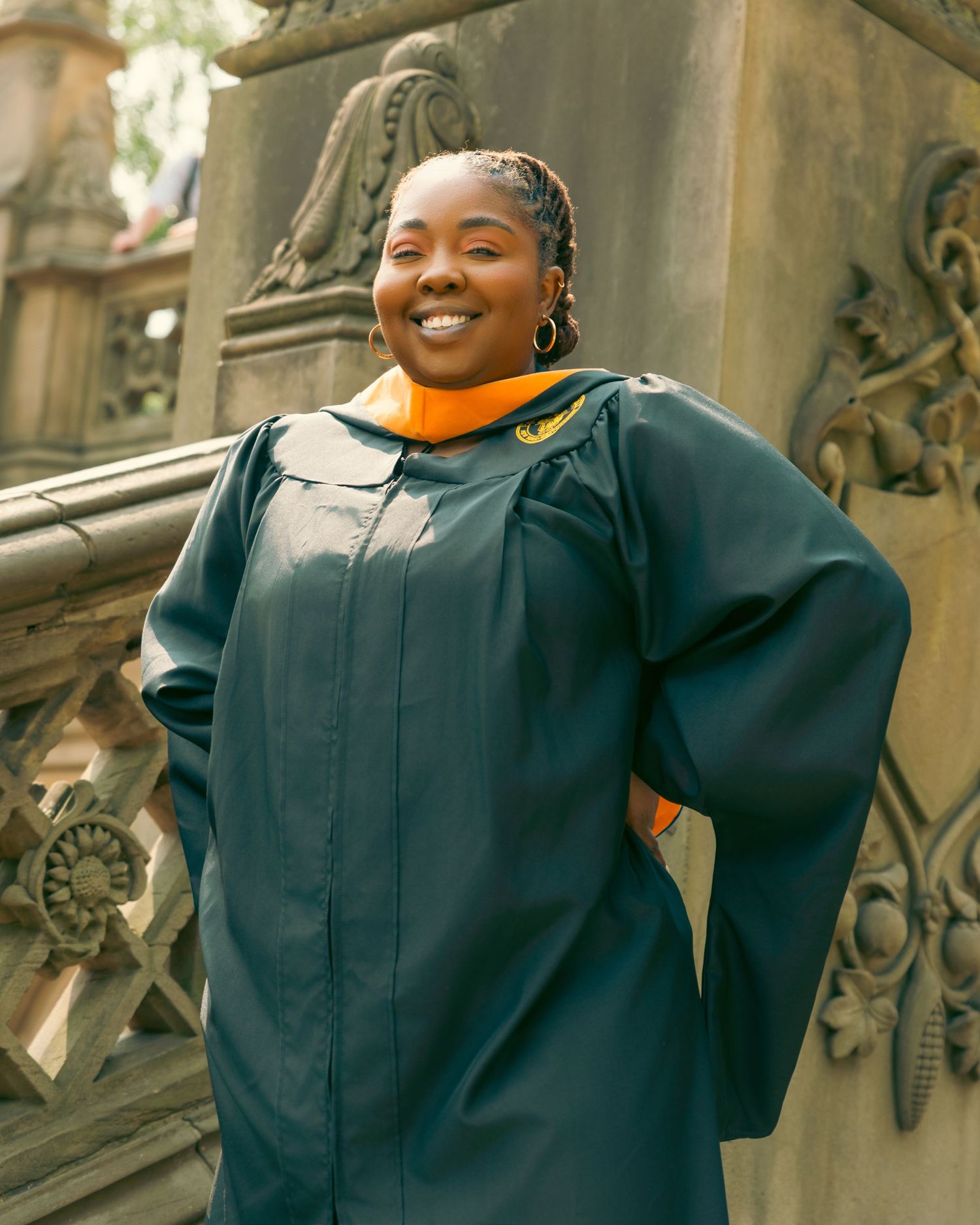Smiling graduate in dark green and gold academic regalia poses by ornate stone architecture.