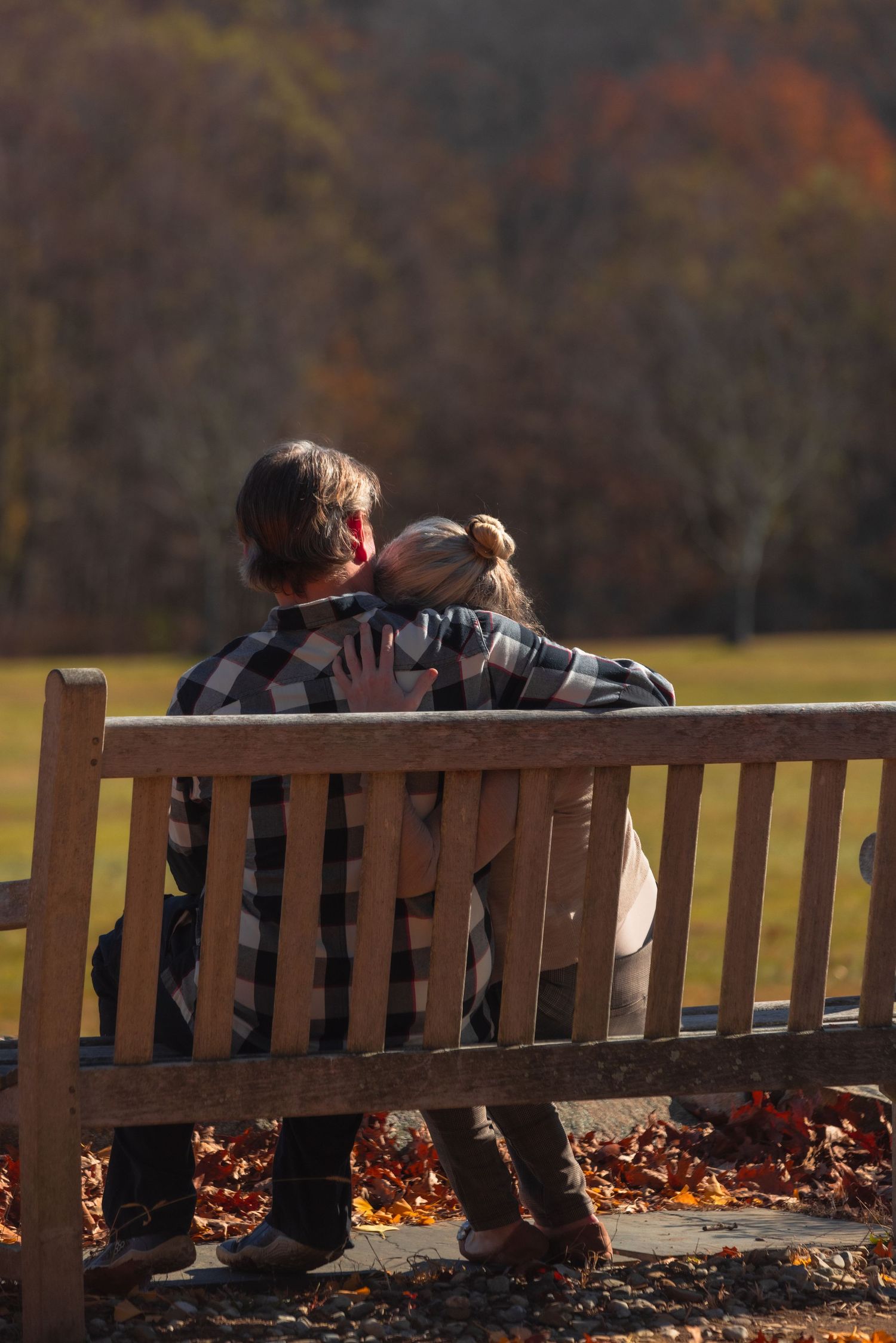 Couple sitting on a wooden bench embracing in an autumn park with colorful fall foliage in the background.