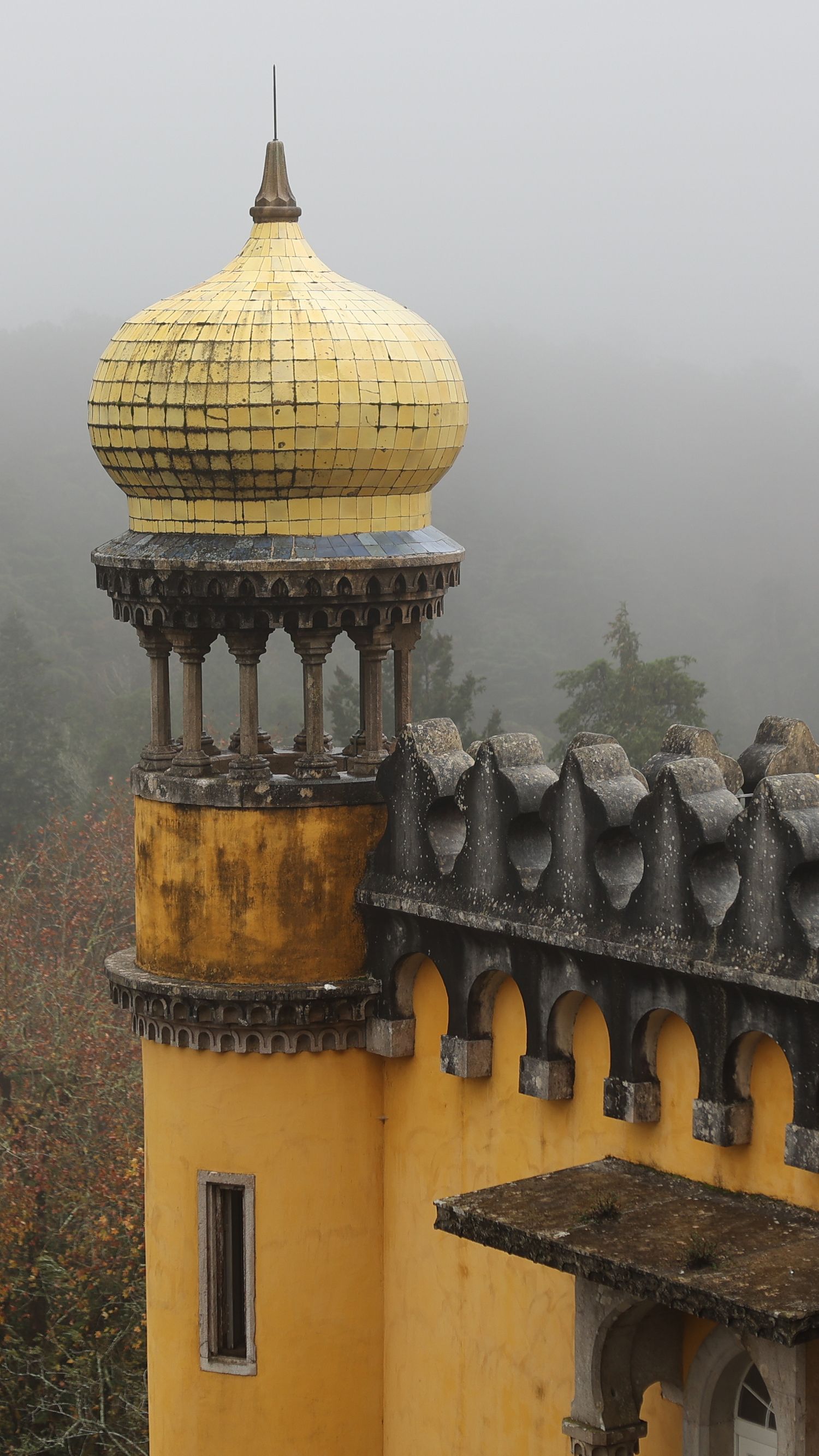 Ornate yellow tiled dome of Pena Palace rising above battlements in misty Sintra, Portugal.