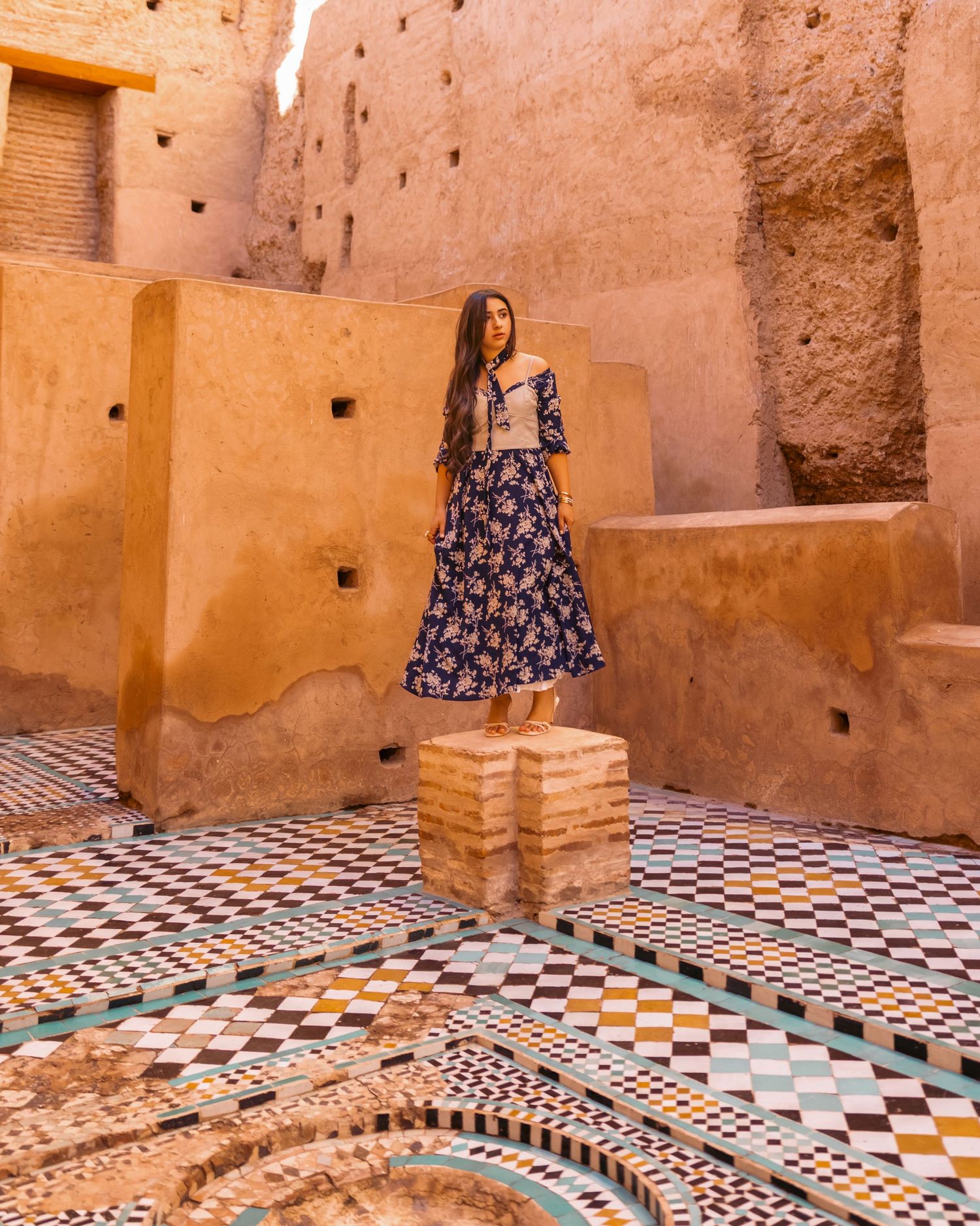 Woman in navy floral dress standing on decorative mosaic tiles in historic Moroccan ruins.