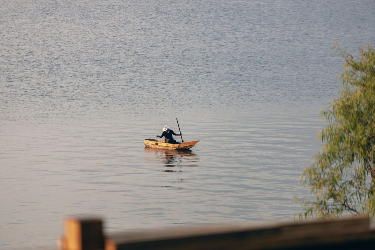 Lone fisherman paddling a small wooden boat on a calm lake at dusk.