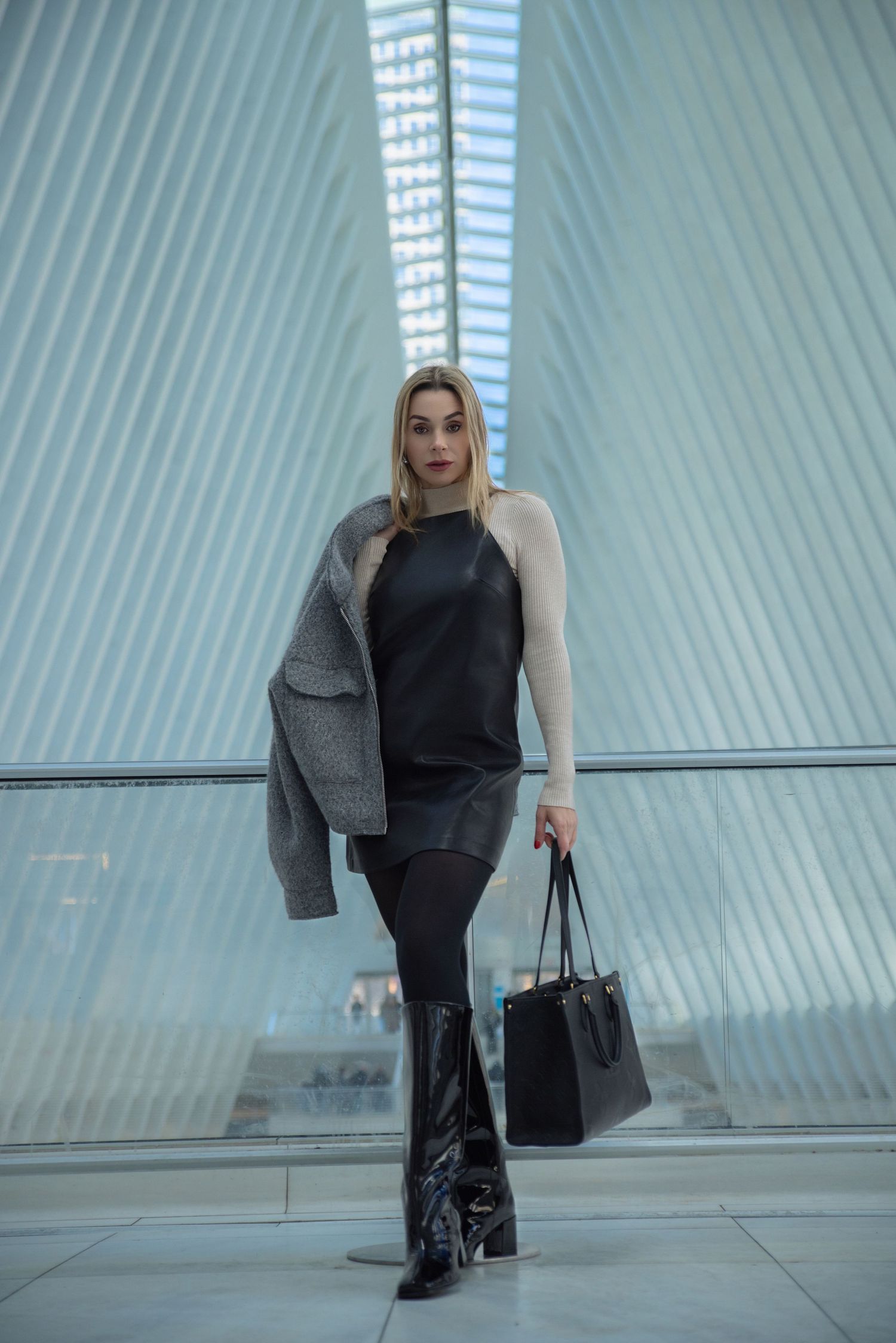 Blonde woman in black leather dress, gray coat, and black boots holding a tote bag in a modern interior.