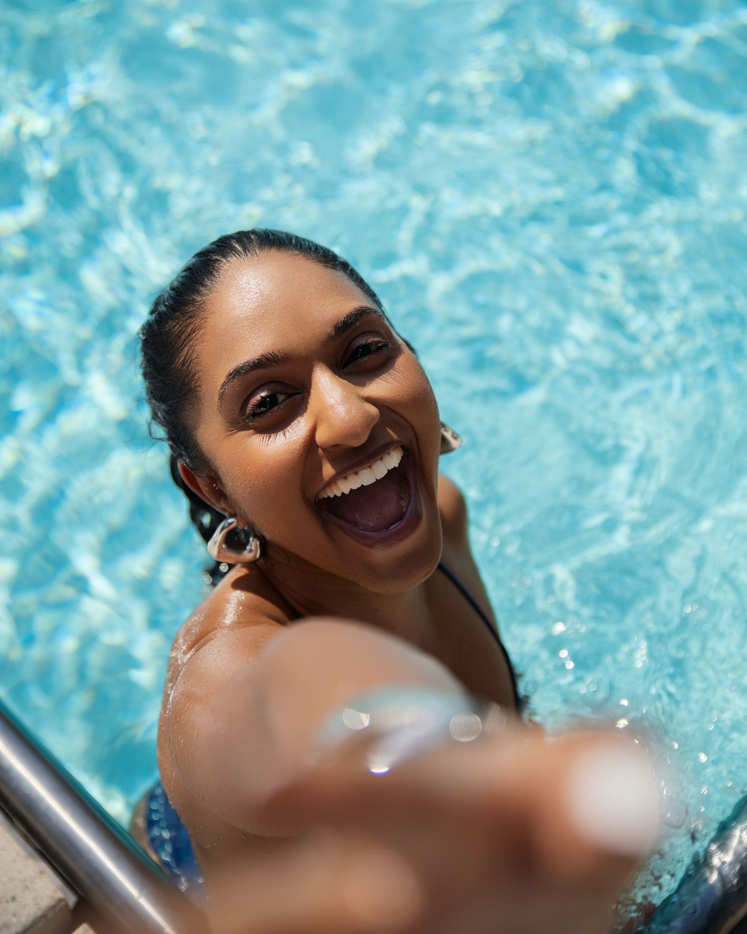 Smiling woman in pool reaching toward camera with bright blue water background on sunny day.