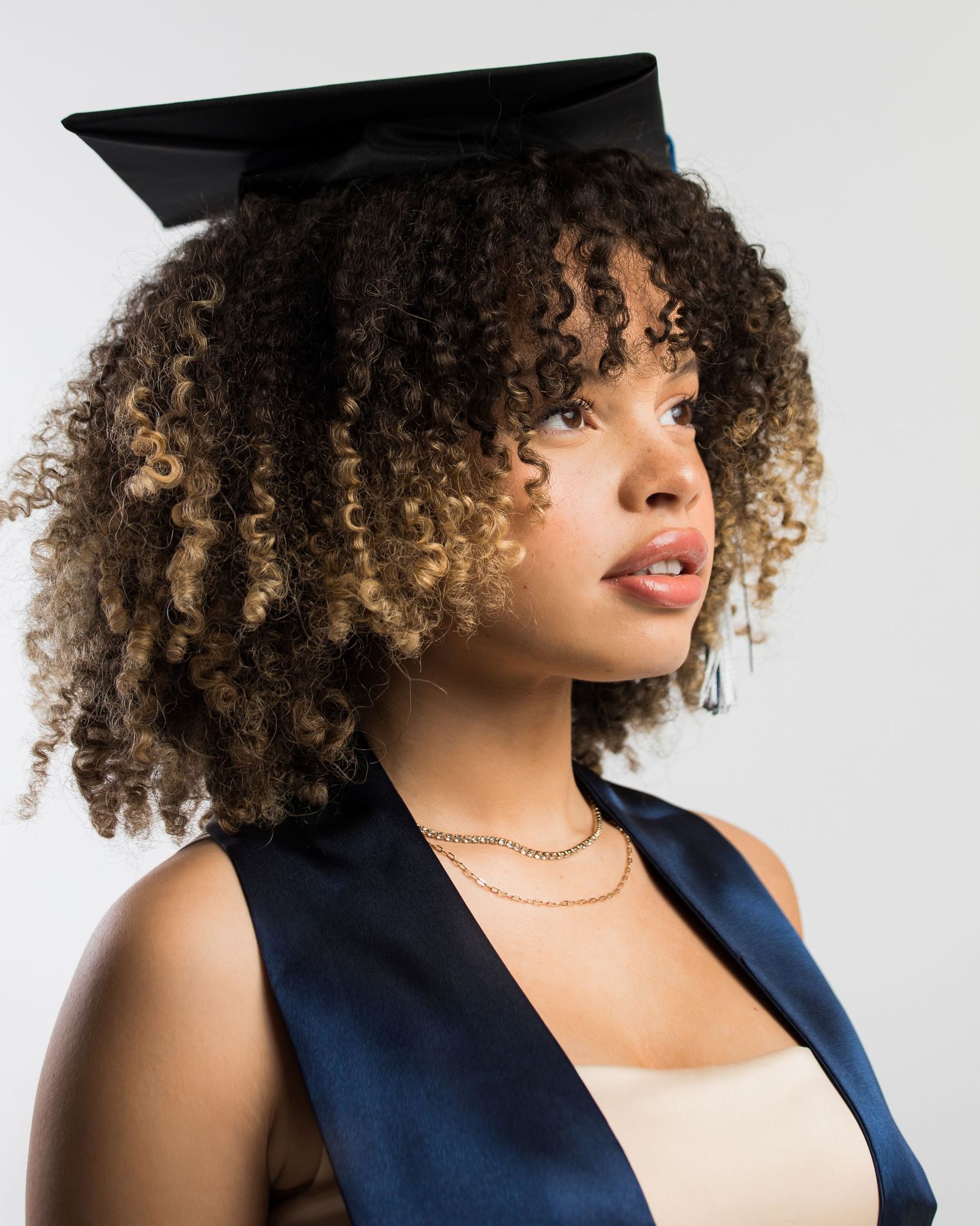Young woman with curly highlighted hair wearing a black graduation cap indoors.