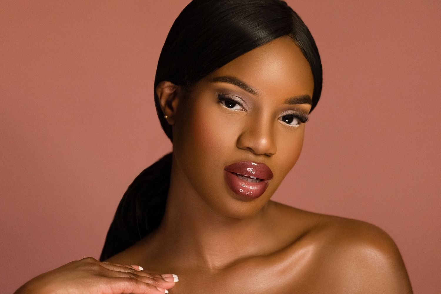 Beautiful woman with sleek hair and glossy lips posing against a warm rose-toned studio background.