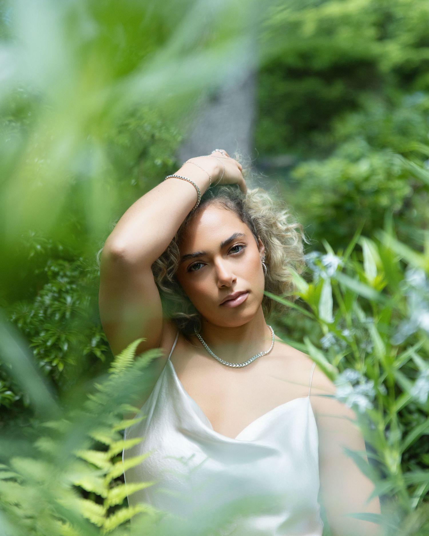 Woman with curly hair in white satin slip dress posing among lush green ferns and garden foliage.