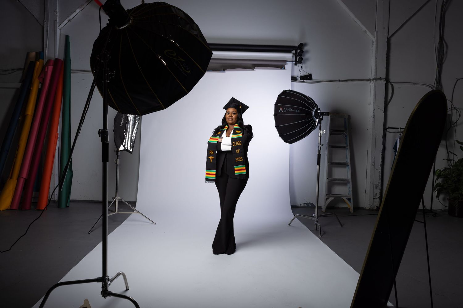 Graduate in cap and gown posing in a professional photo studio with softbox lighting equipment visible.