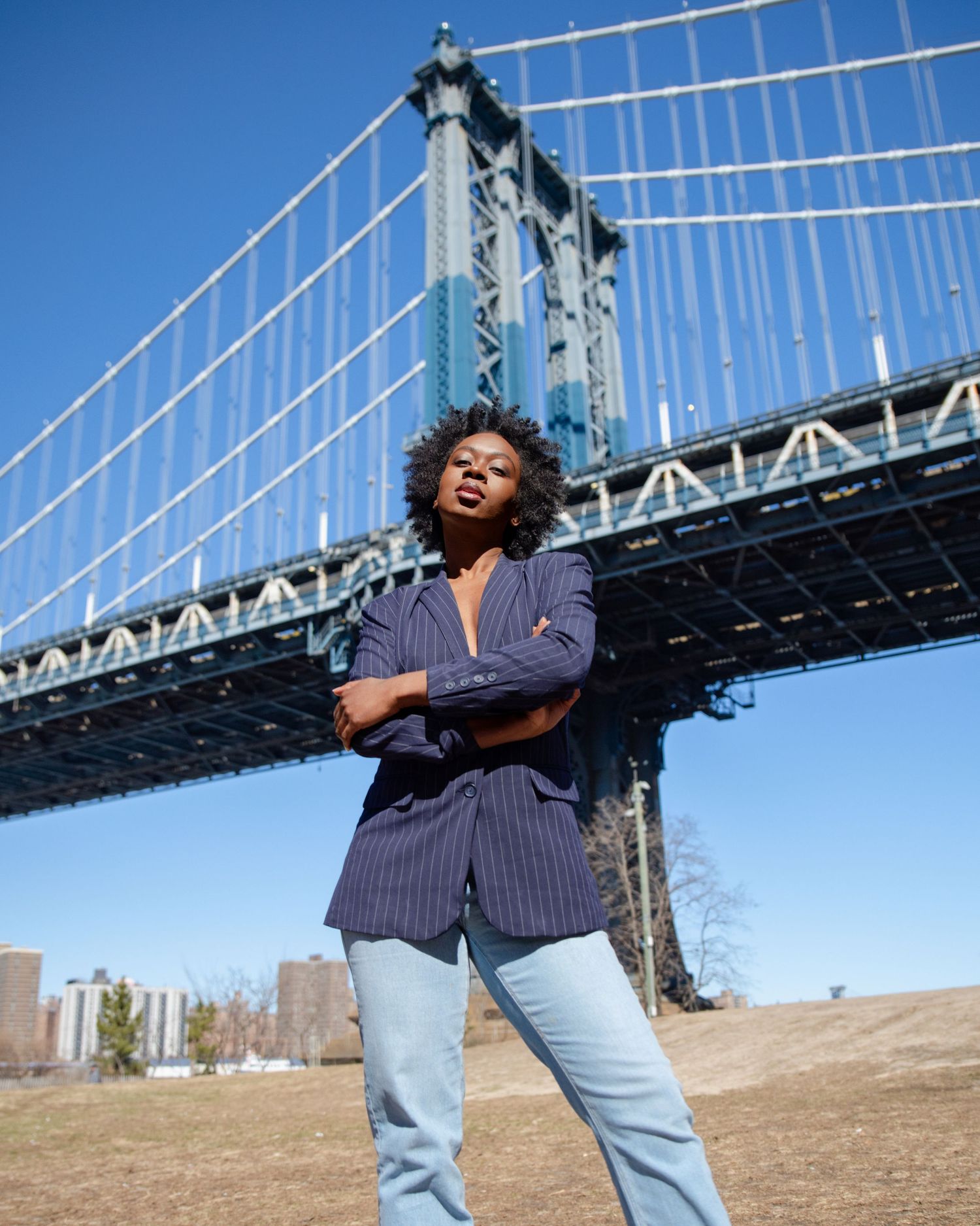 Woman in navy pinstripe blazer and jeans posing confidently beneath the Manhattan Bridge on a sunny day.