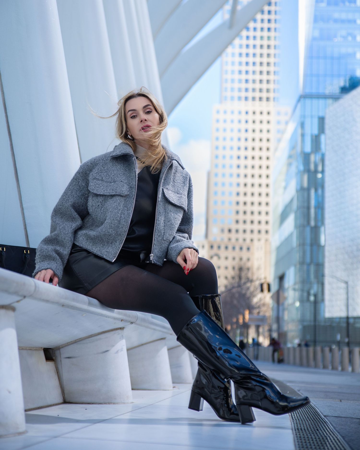 Blonde woman in gray jacket, black dress, and knee-high boots sitting on white steps in city.