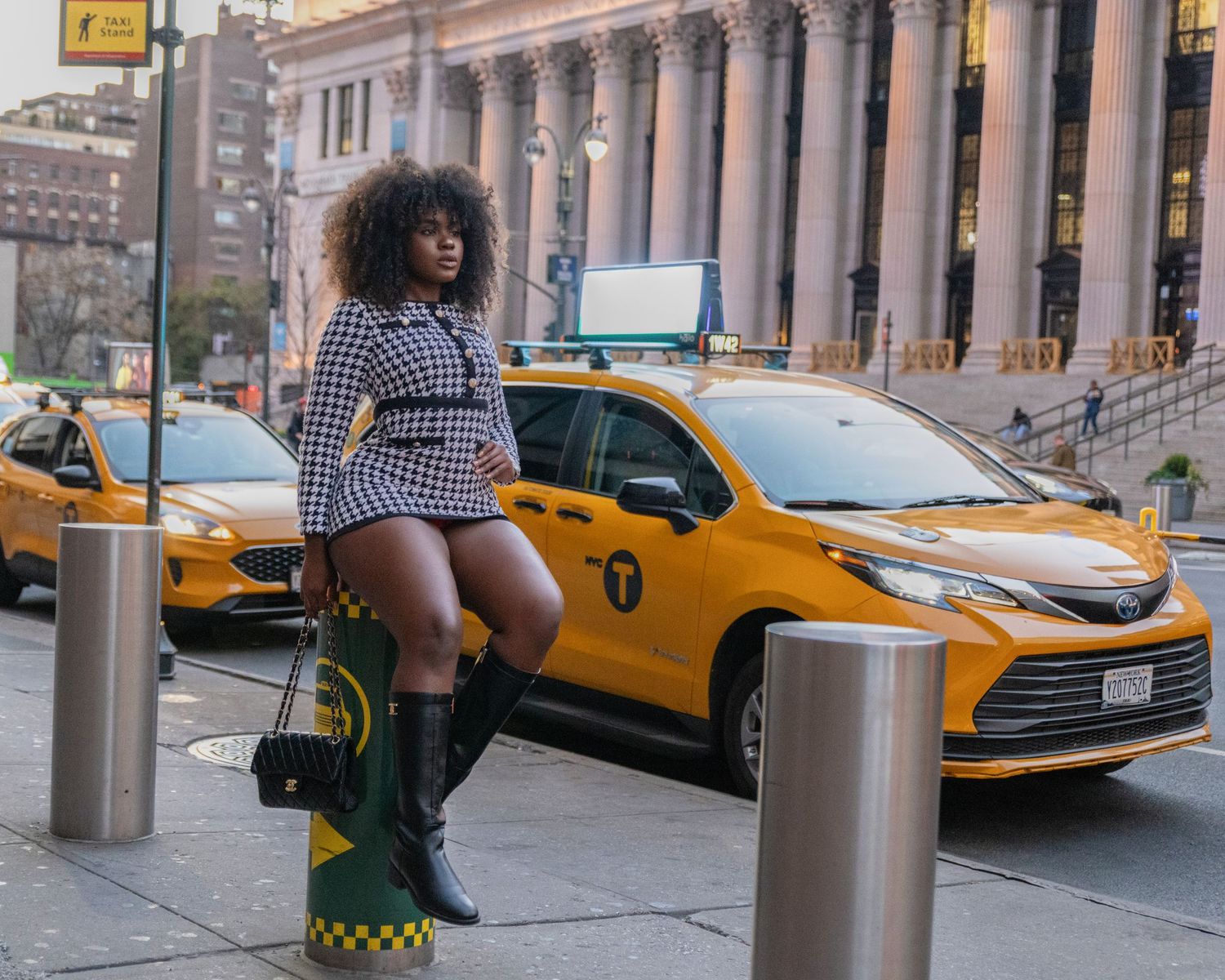 Curvy woman in houndstooth mini skirt and black boots posing near yellow taxis on NYC street.