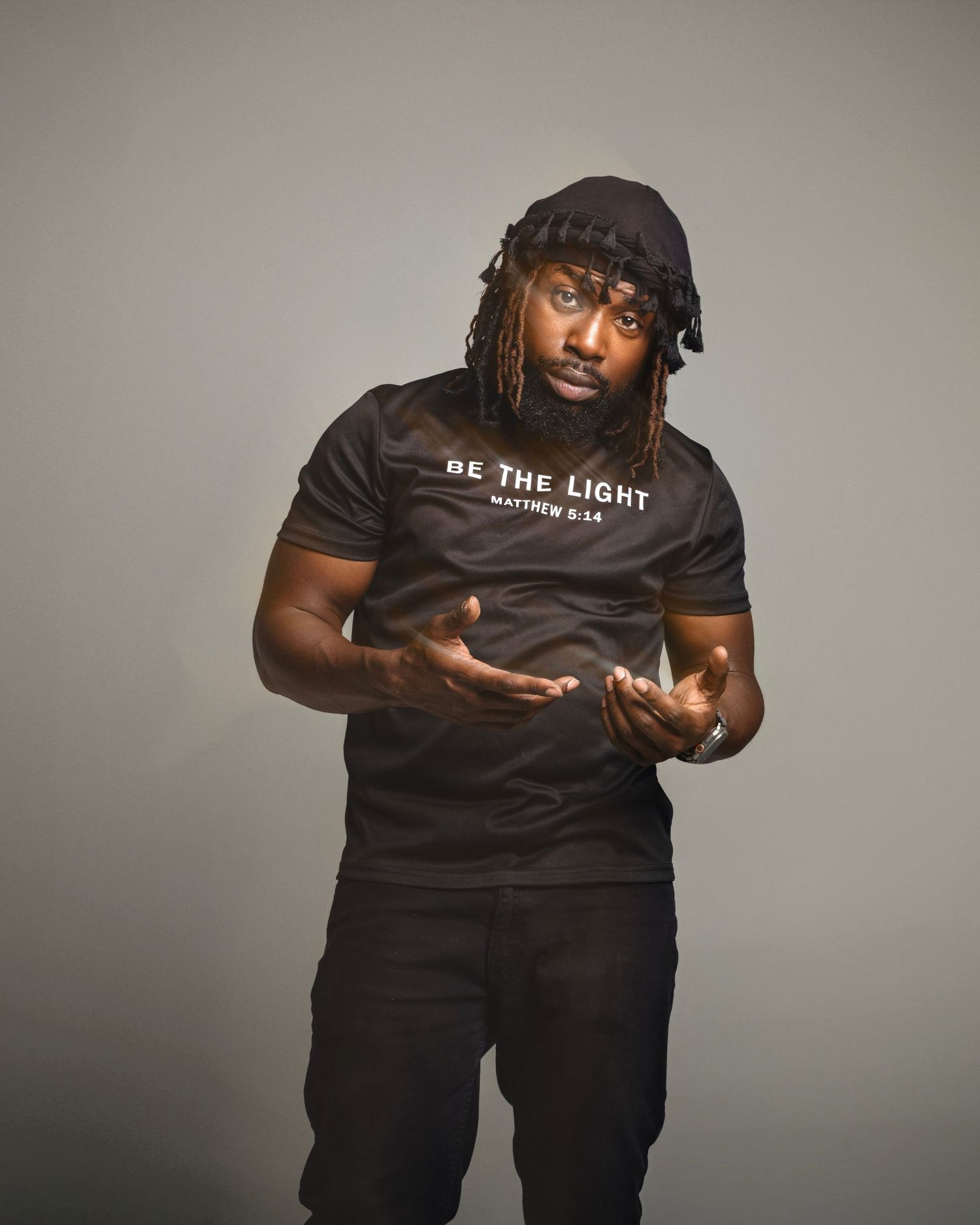 Man in black Be The Light Matthew 5:14 t-shirt with dreadlocks gesturing with hands against gray background.