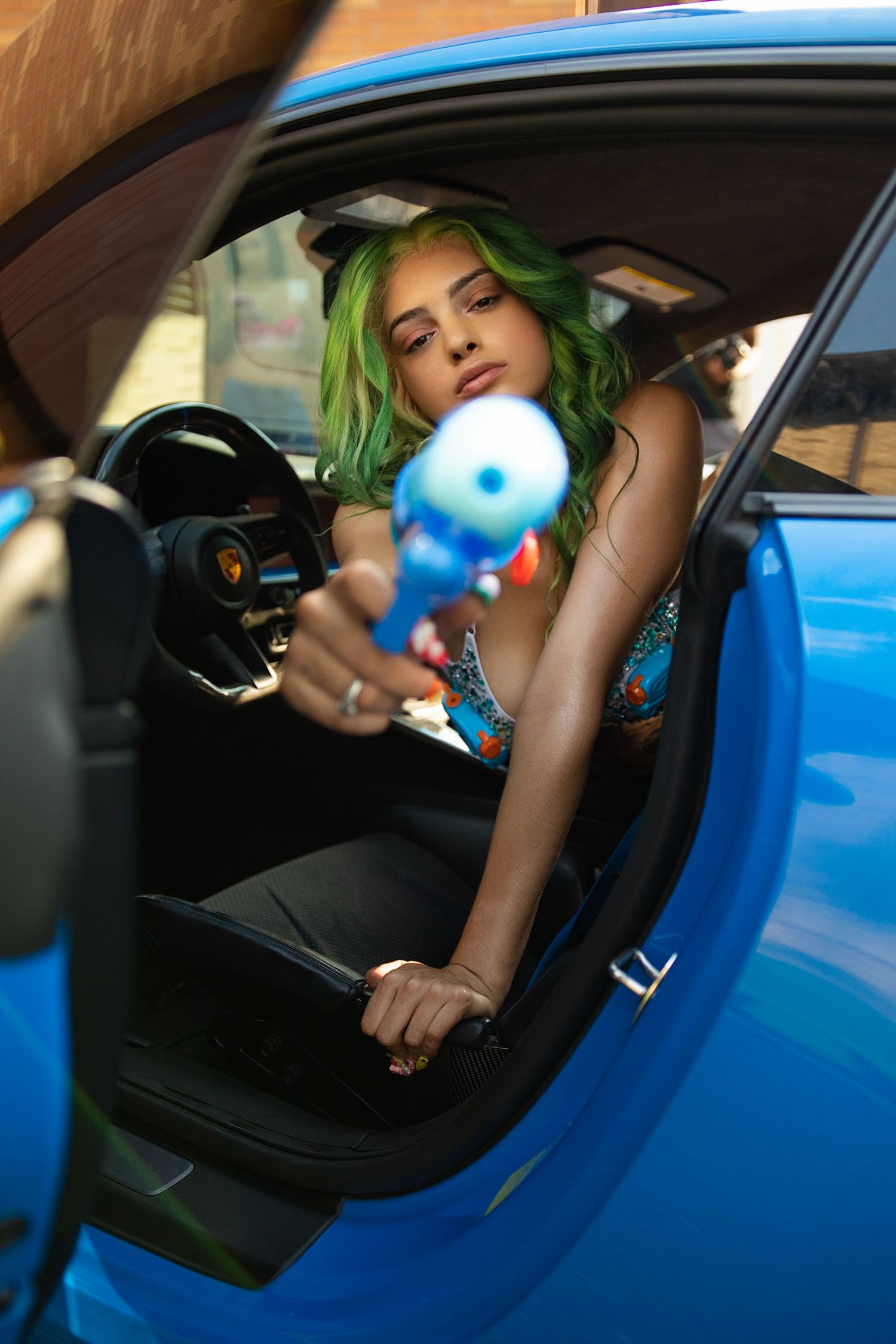 Green-haired woman pointing blue toy gun from blue Porsche sports car interior.