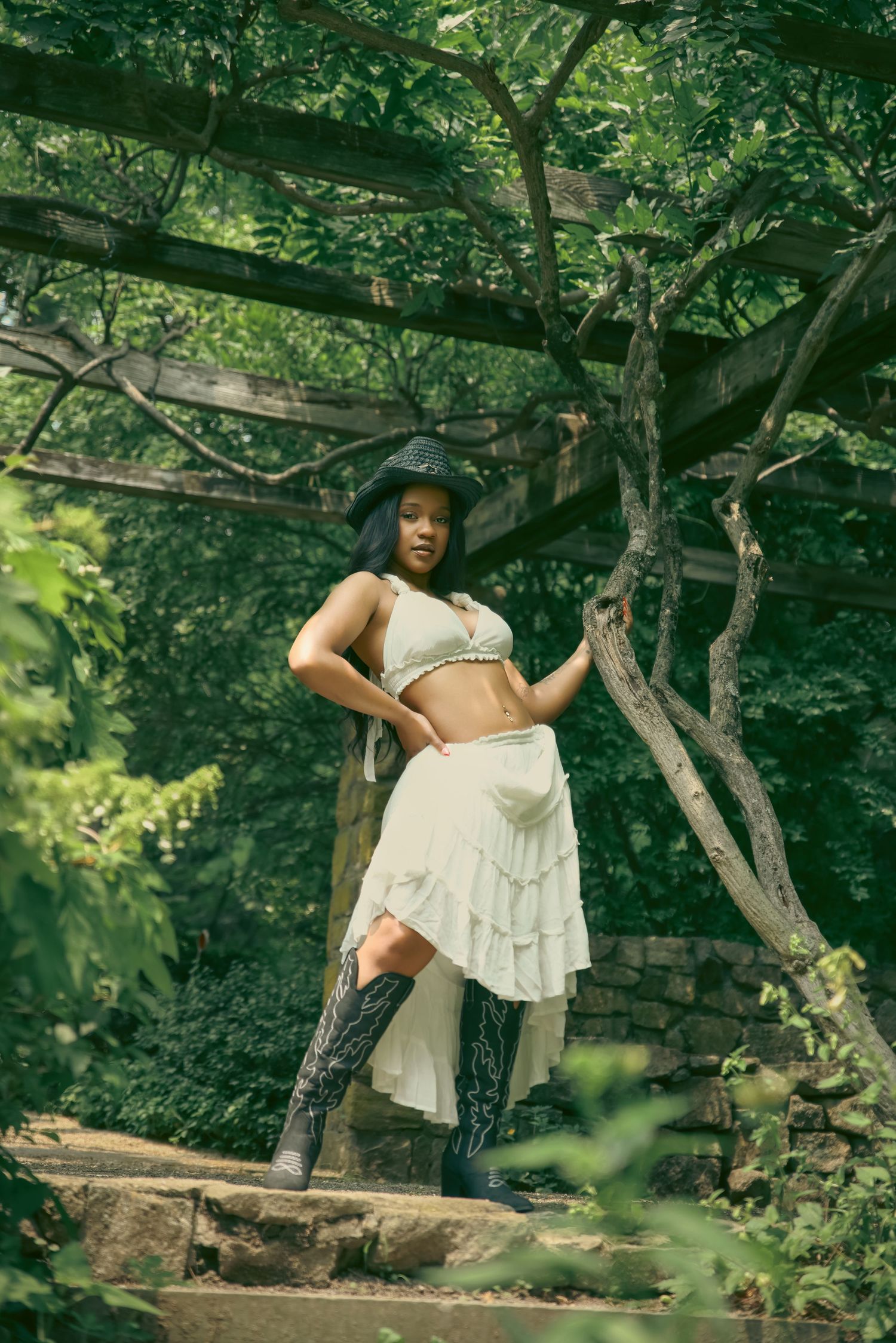 Woman in white crop top, ruffled skirt, cowboy boots, and black hat posing under a vine-covered pergola.