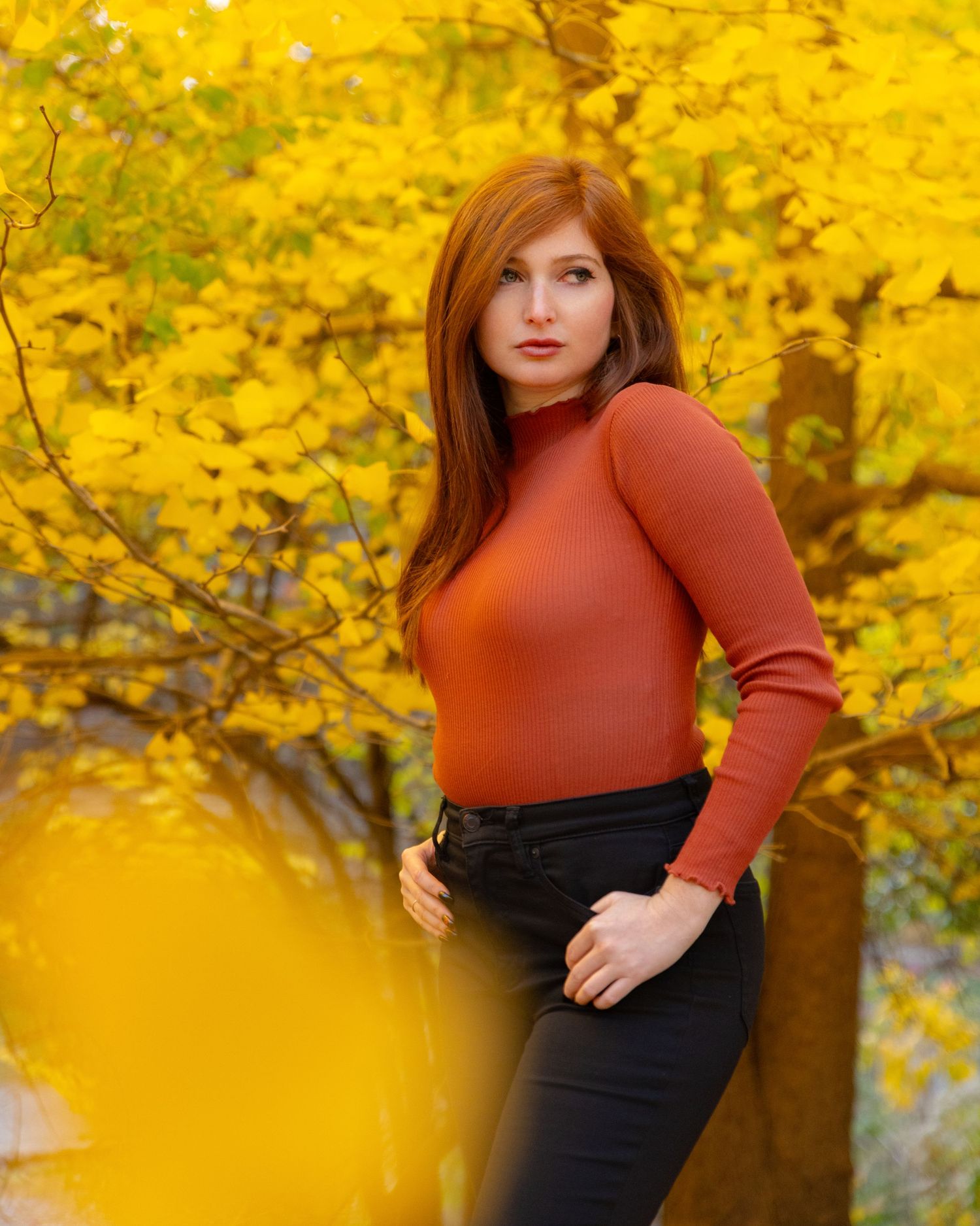 Woman in rust turtleneck and black jeans poses among vibrant yellow autumn foliage.