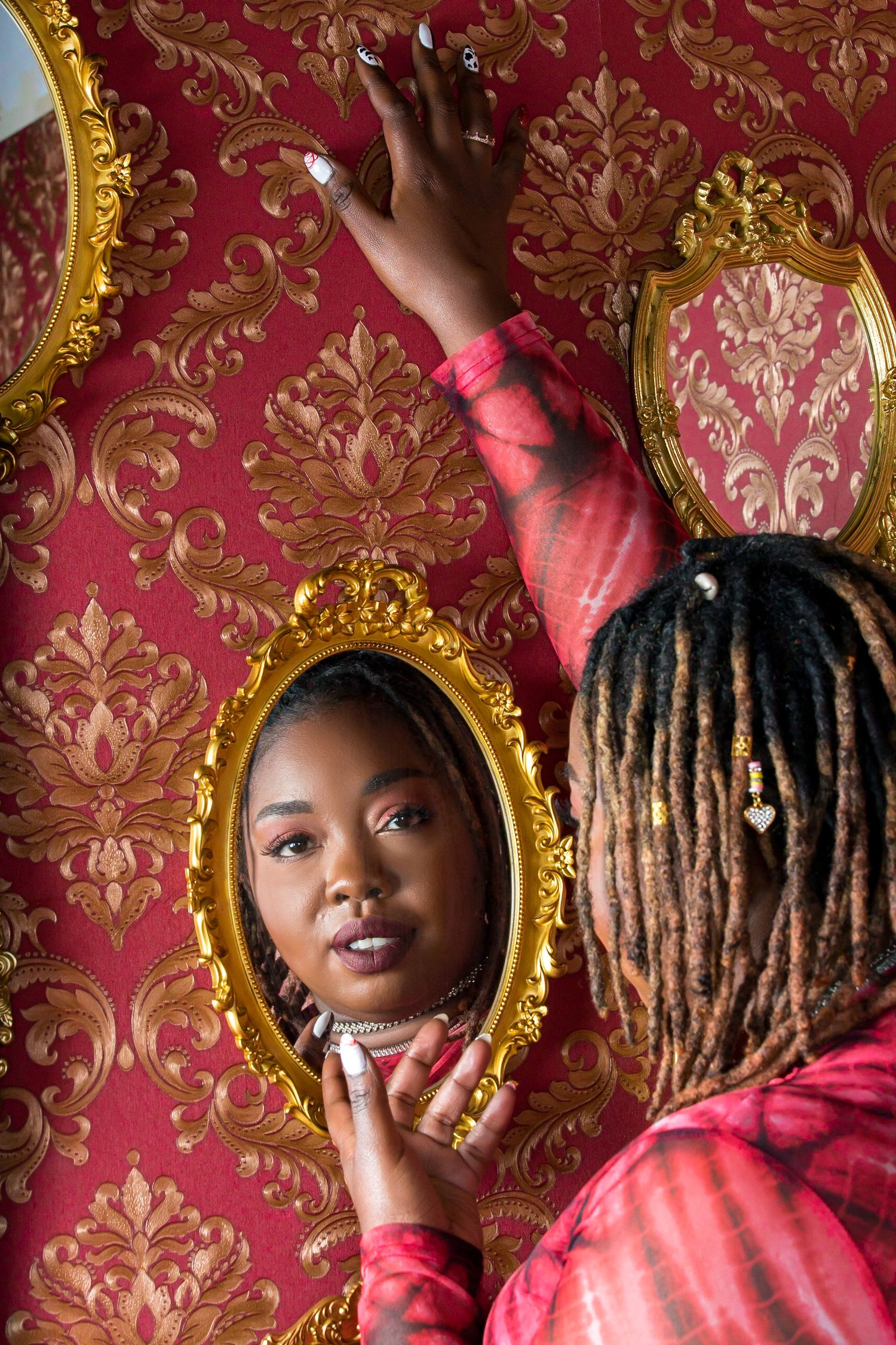 Woman with locs views reflection in ornate gold mirror on red damask wallpaper.