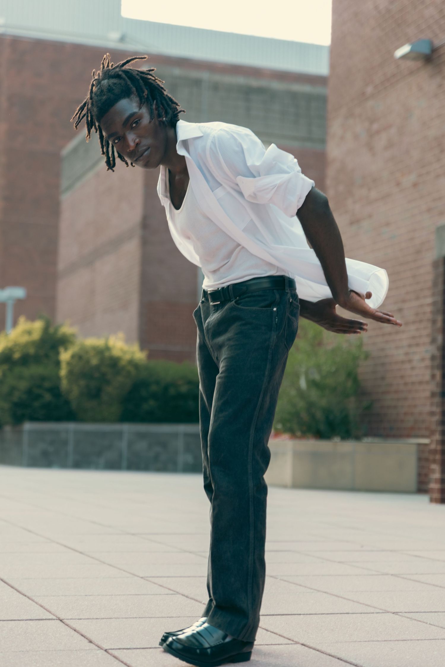 Young Black man with dreadlocks in white shirt and dark jeans posing outdoors near brick building.