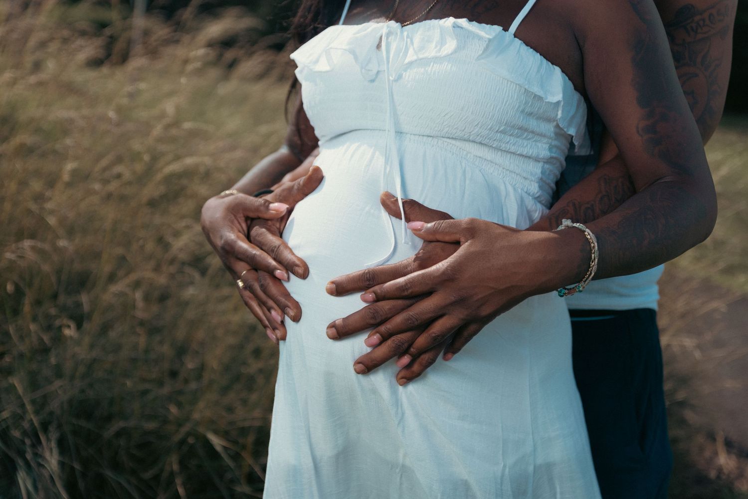 Close-up of pregnant woman in white dress with partner's hands cradling baby bump outdoors.