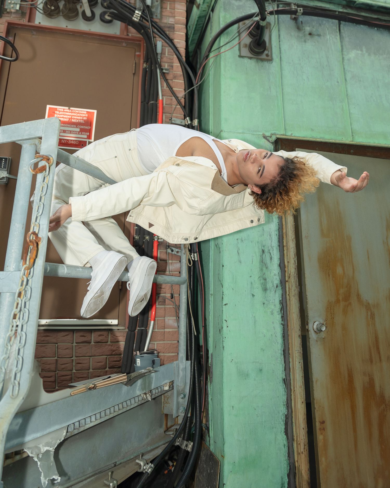 Person in white outfit performing dynamic pose on industrial rooftop fire escape structure.