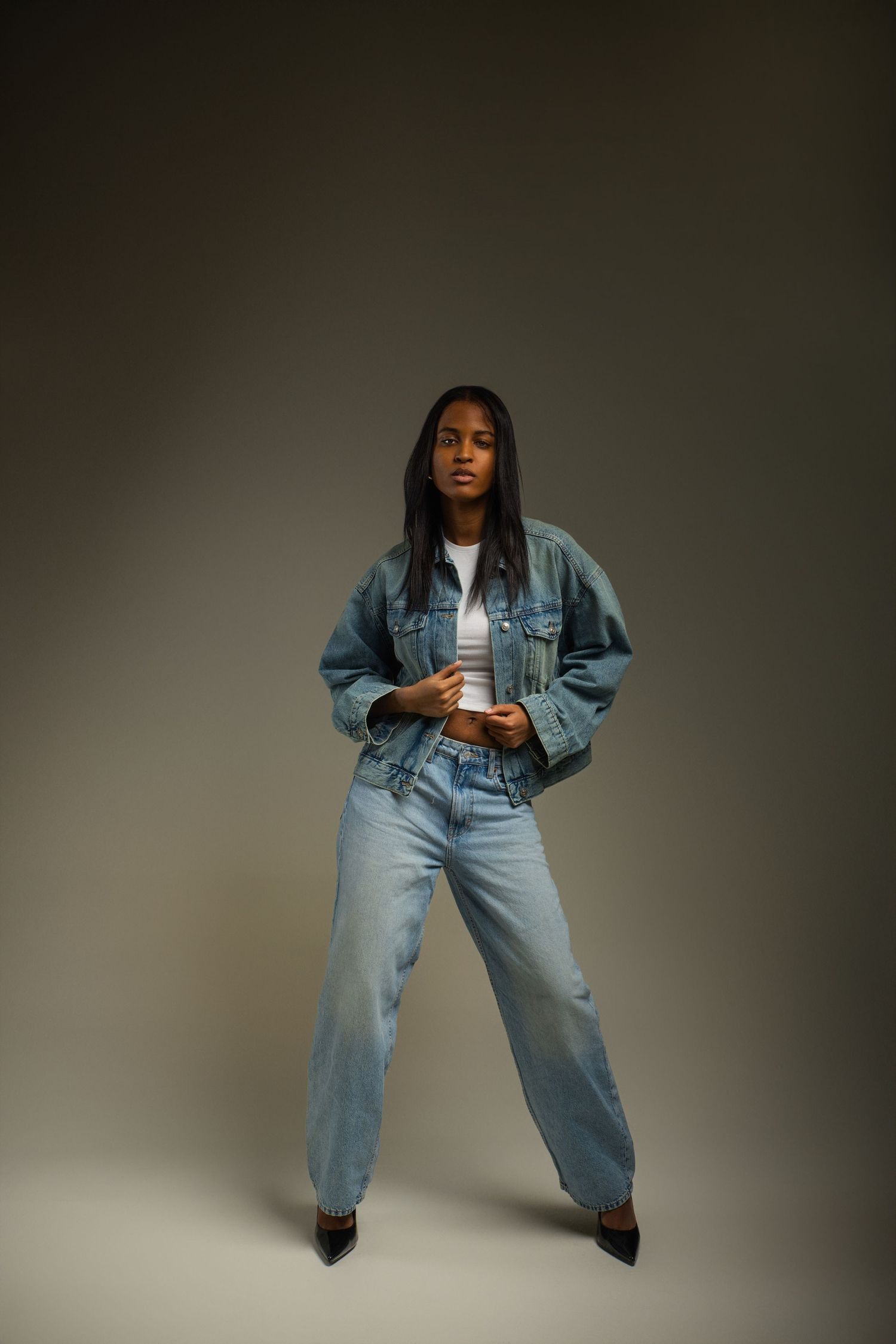 Young Black woman in denim jacket and wide-leg jeans posing confidently in studio with neutral background.