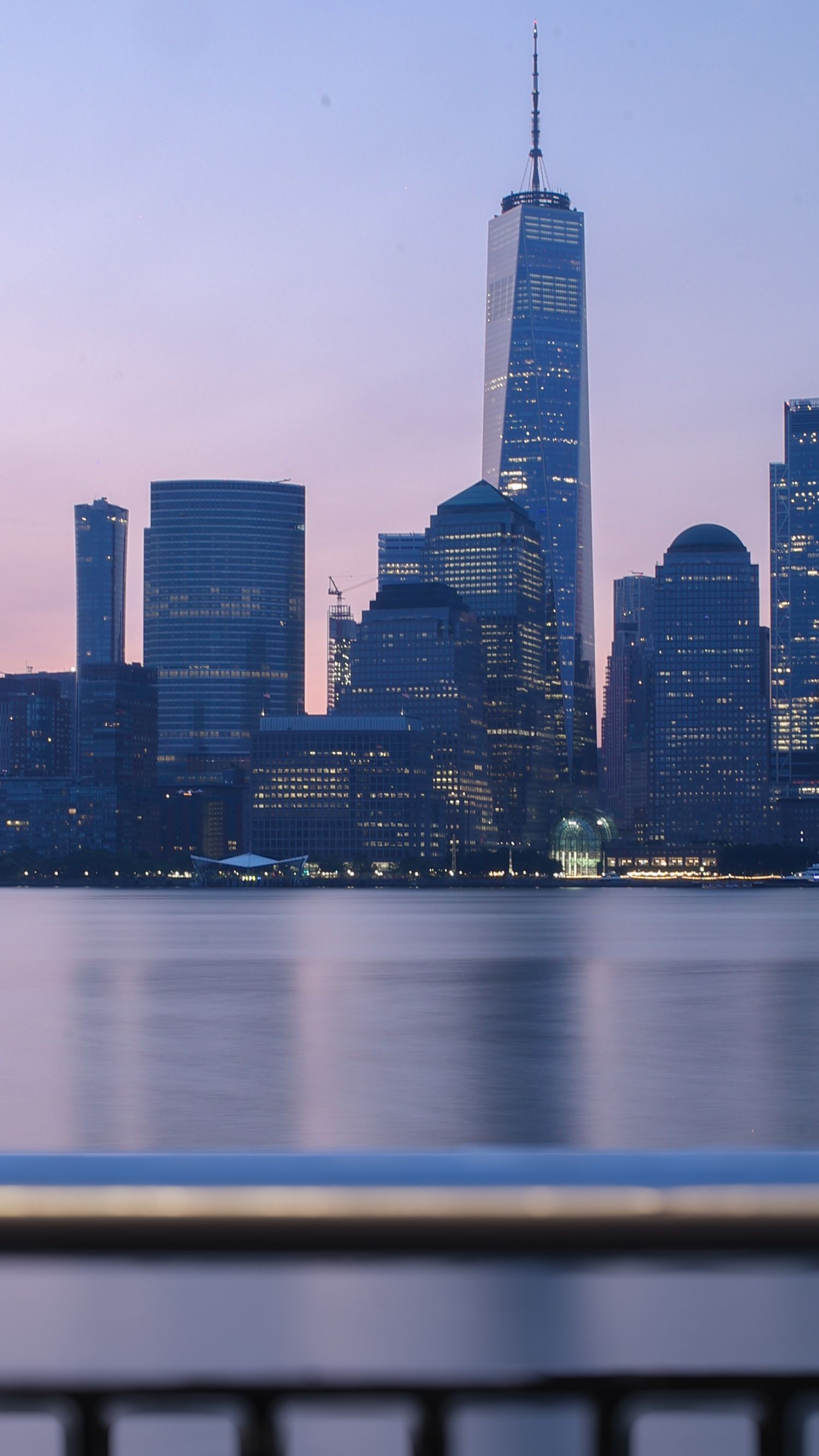 One World Trade Center and Manhattan skyline reflecting on calm Hudson River water at dusk with pink sky.