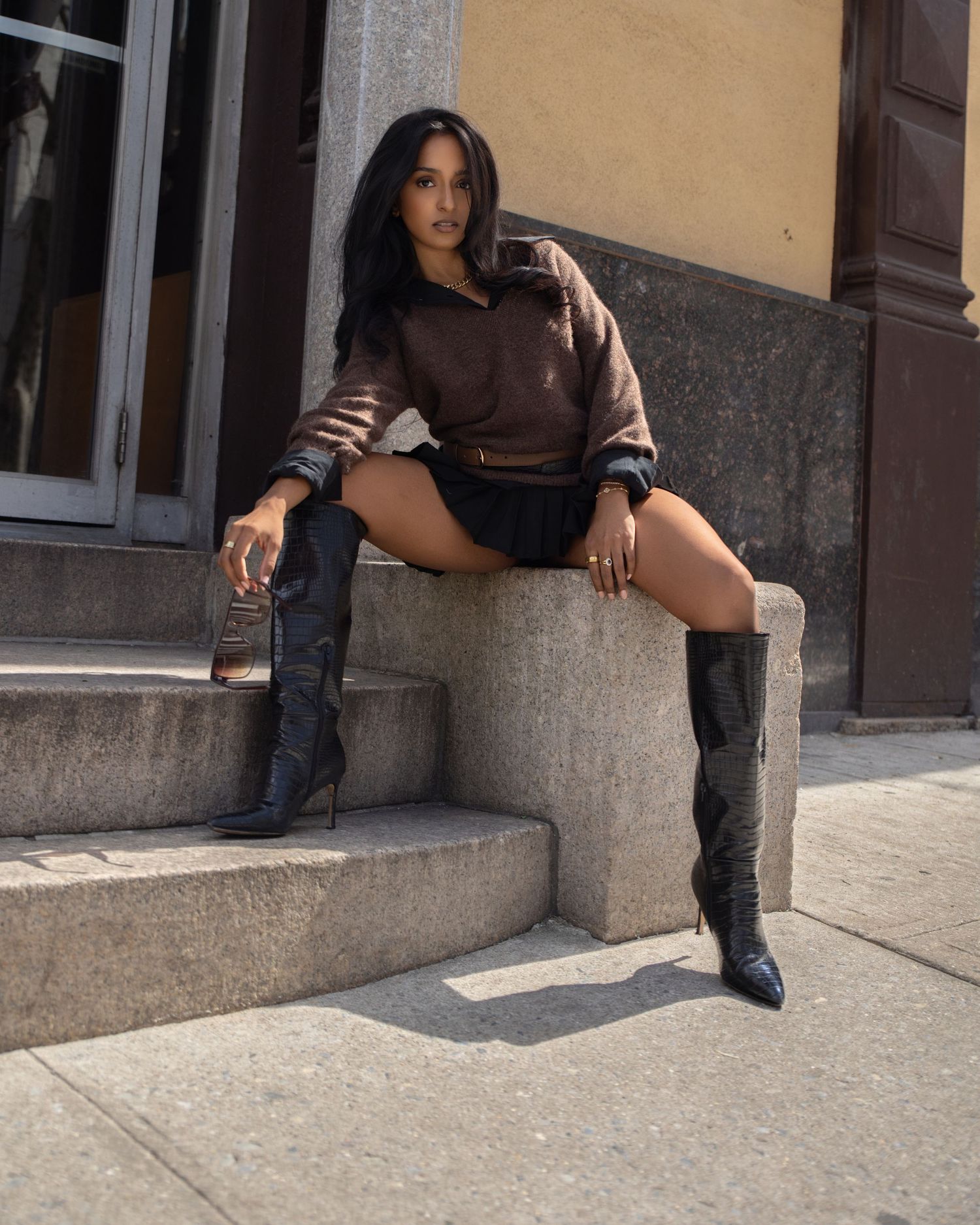 Stylish woman in brown cropped hoodie and black knee-high boots posing on stone steps outside a building.