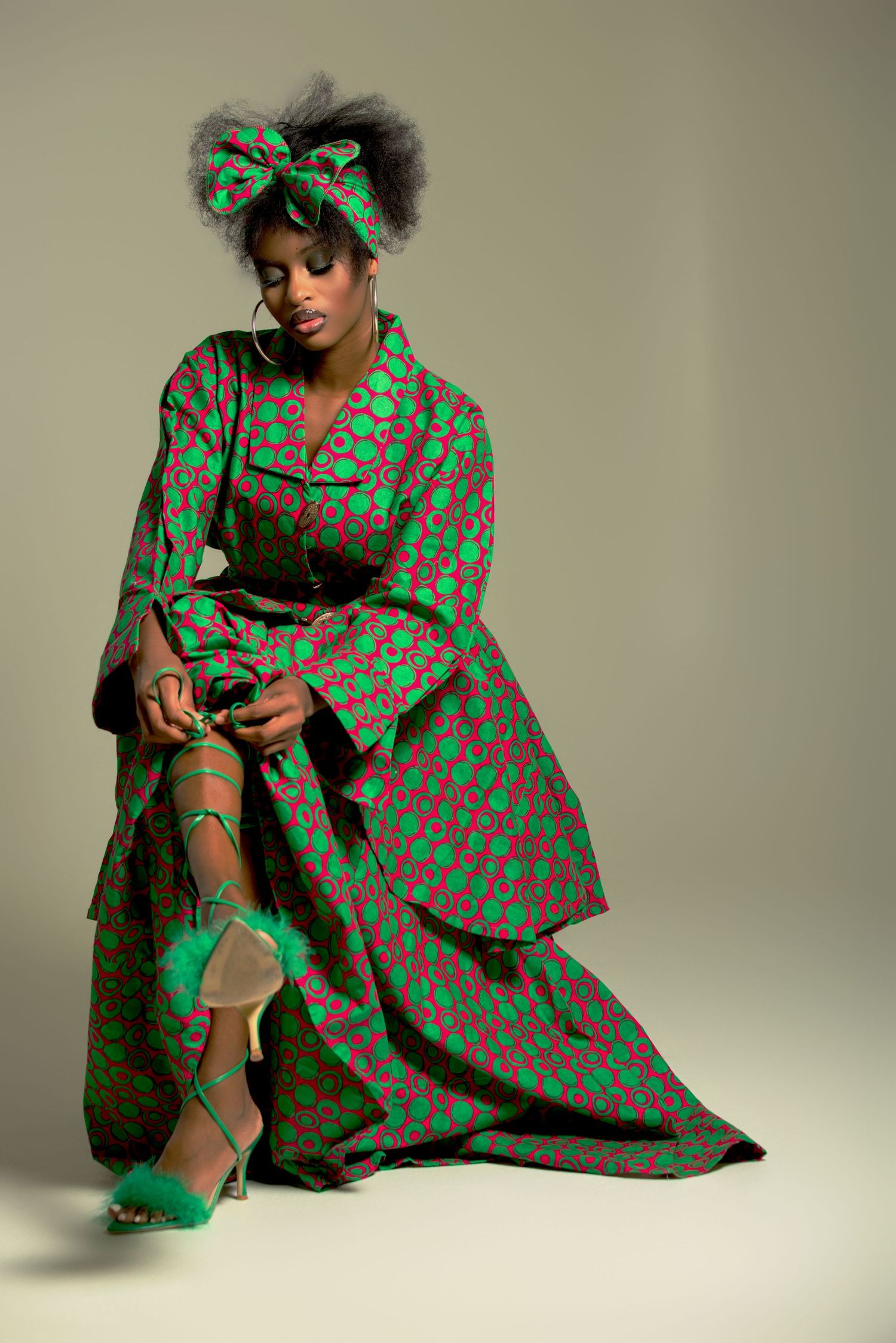 Woman in green and red African print dress with matching headwrap, seated gracefully in a studio setting.
