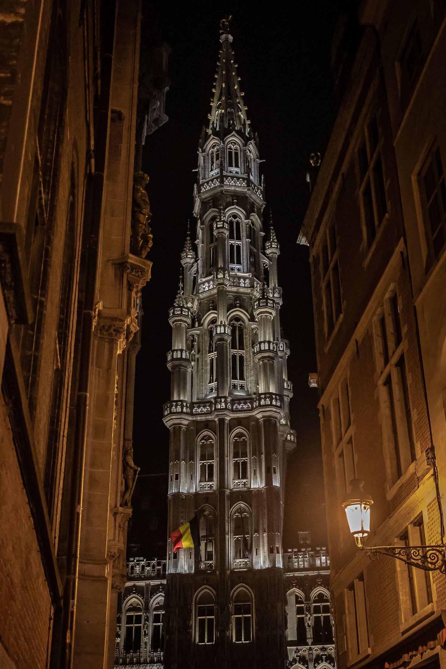 Brussels Town Hall Gothic tower illuminated at night, viewed through a narrow alley with a street lamp.