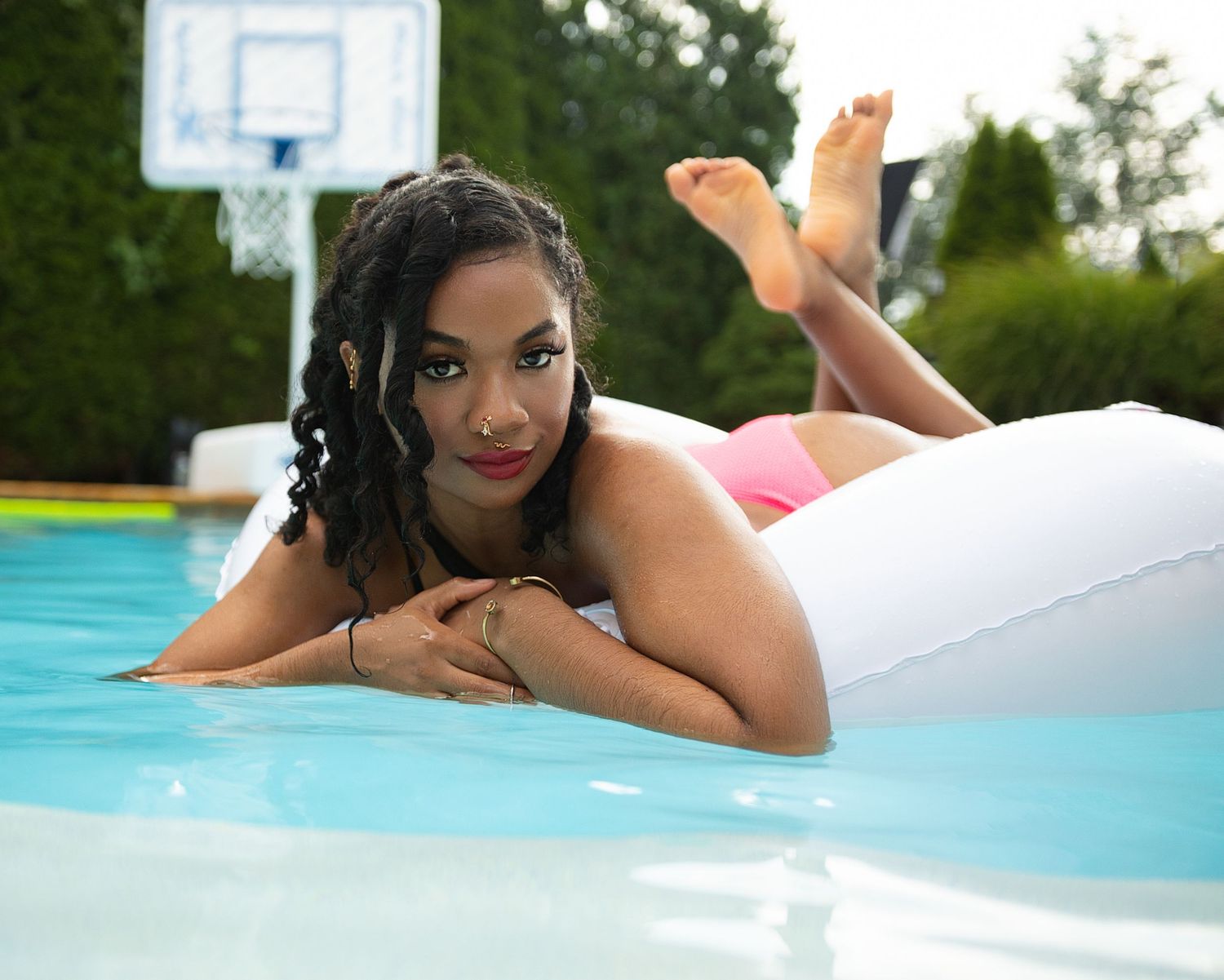 Young woman lying on white pool float in outdoor swimming pool with basketball hoop.