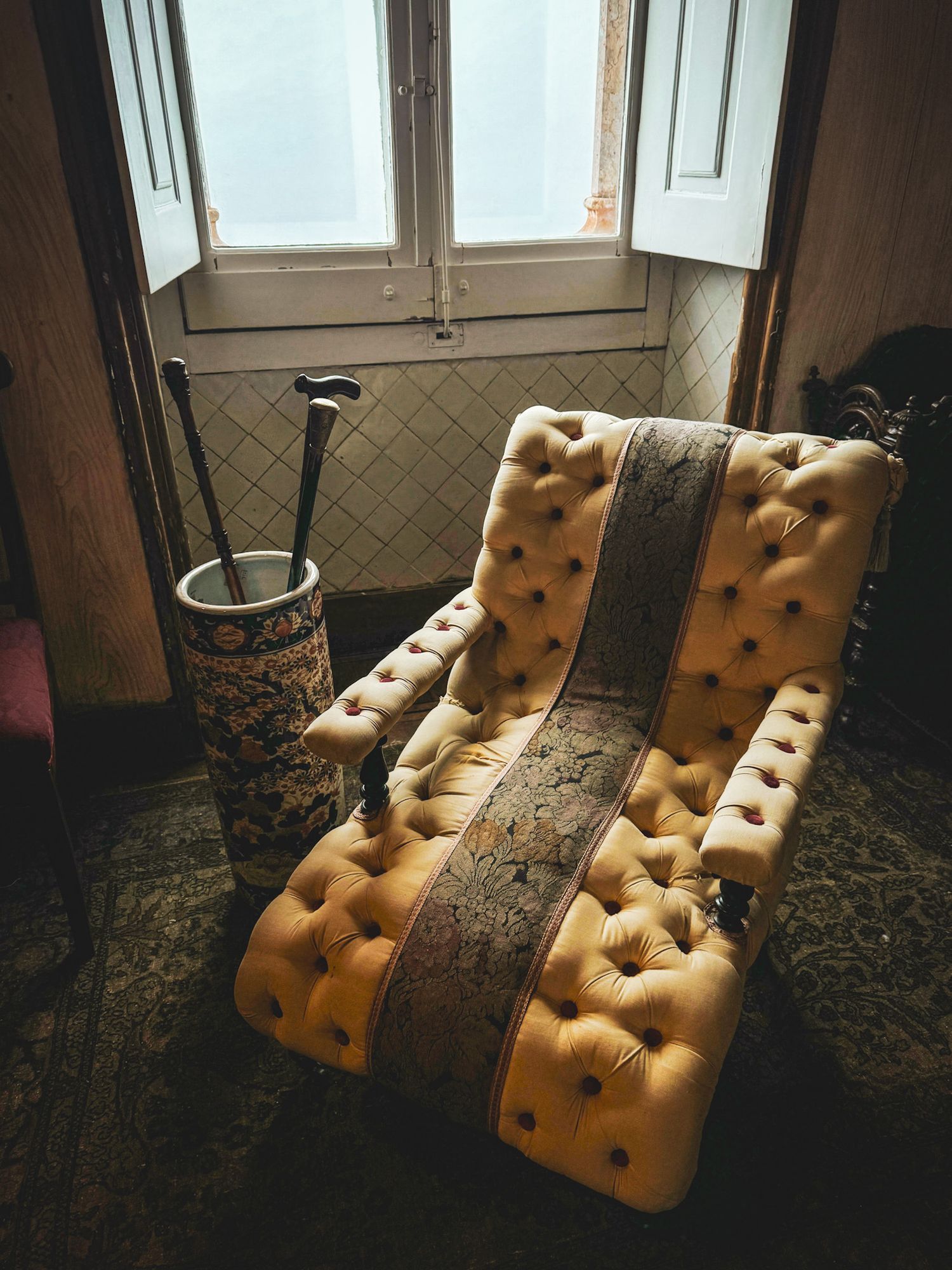 Vintage tufted armchair near a sunlit window with an ornate umbrella stand in a rustic room.