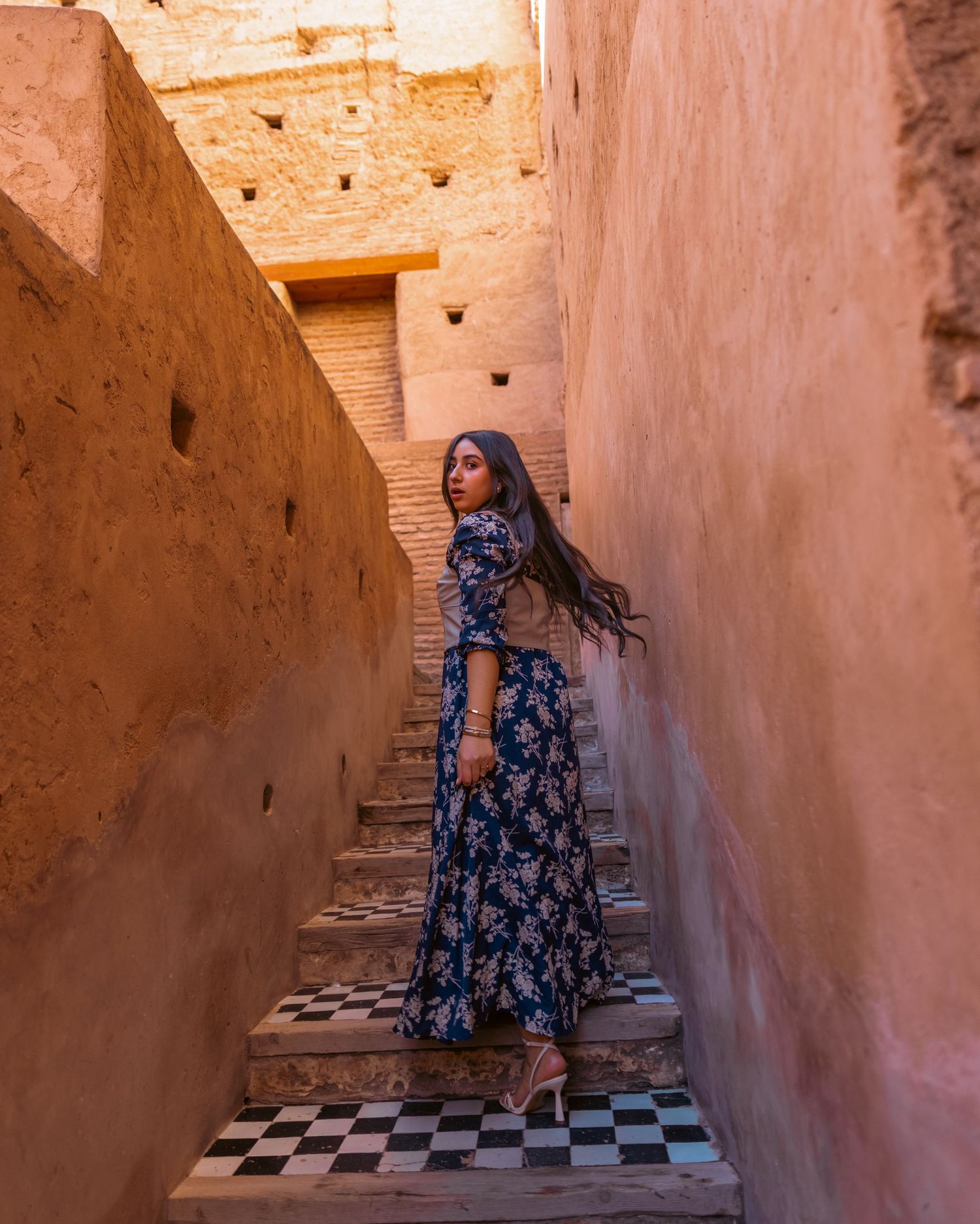 Woman in blue floral maxi dress on tiled stairs in ancient Moroccan architecture.
