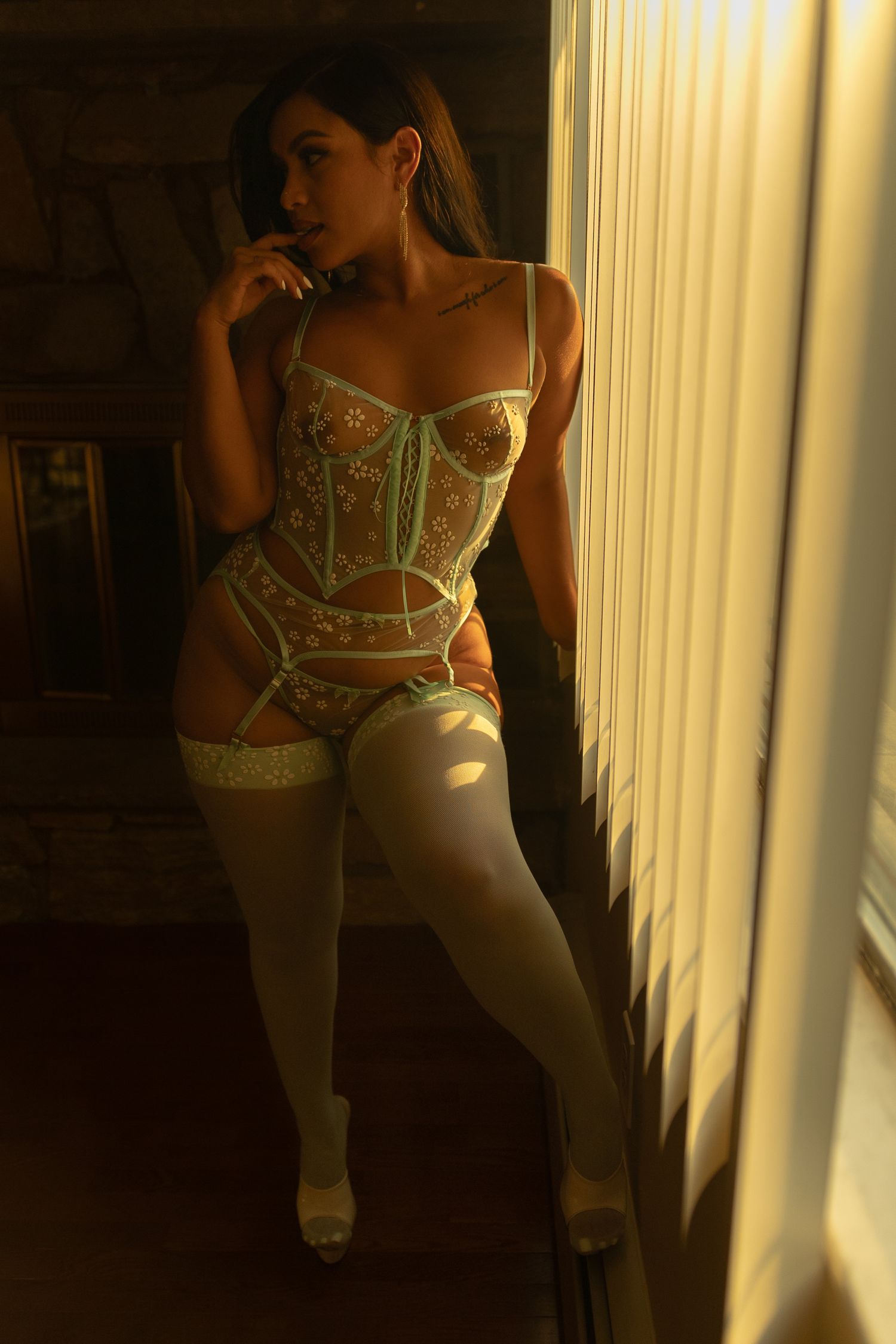 Woman in floral corset lingerie and white stockings standing by window in warm golden light.