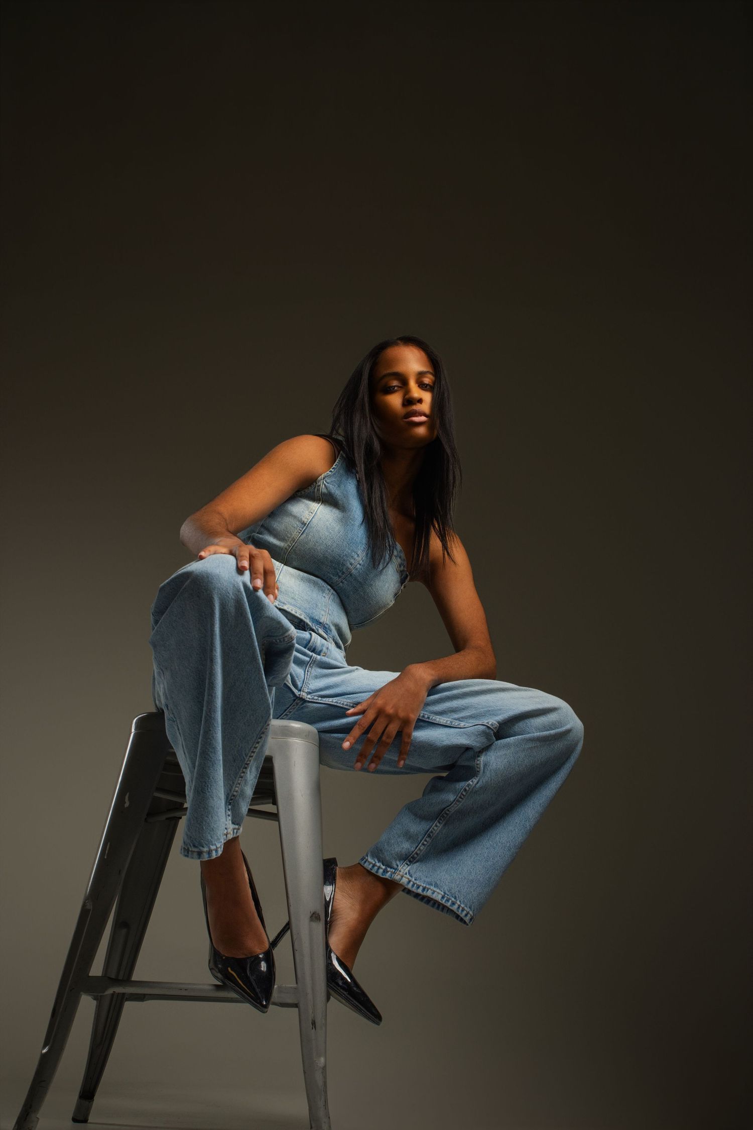Woman in light blue denim jumpsuit sitting on a metal stool with black heels against a dark studio background.