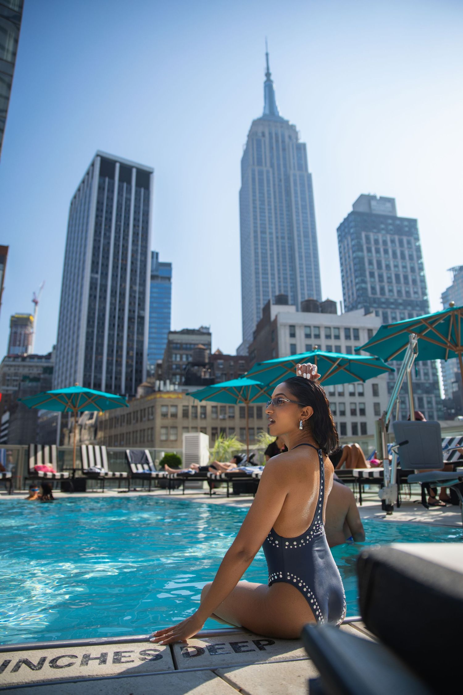 Woman in black swimsuit sitting by rooftop pool with Empire State Building skyline in background.