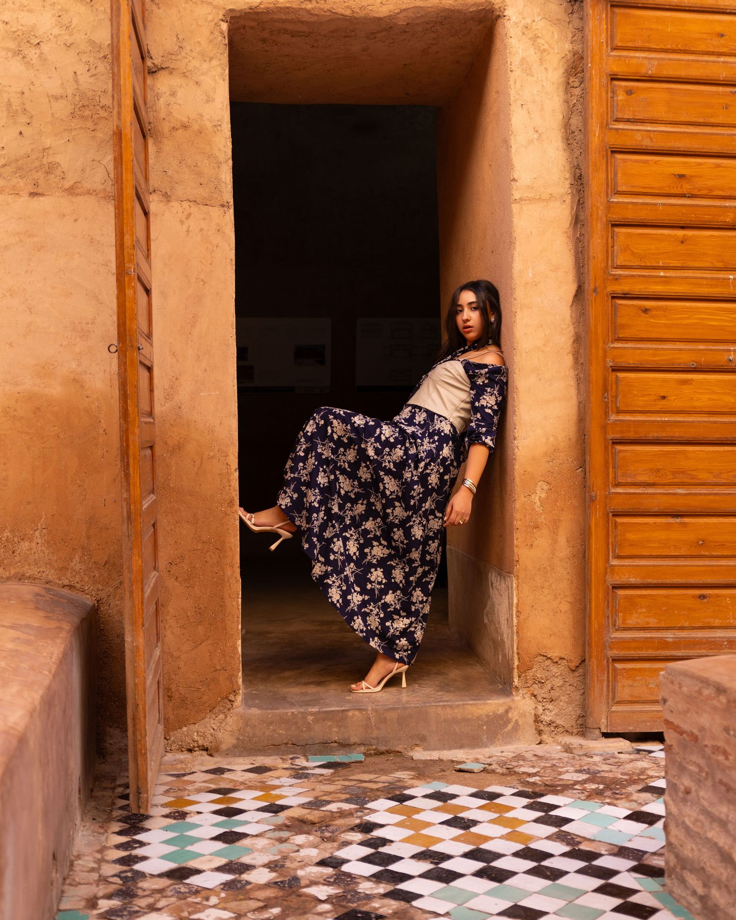 Woman in blue floral skirt posing in ancient Moroccan doorway with checkered tile floor.