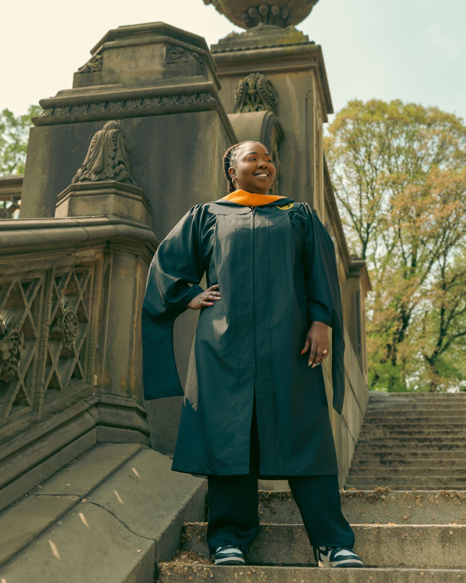 Happy graduate in teal and gold commencement gown stands on stone steps outdoors among trees.